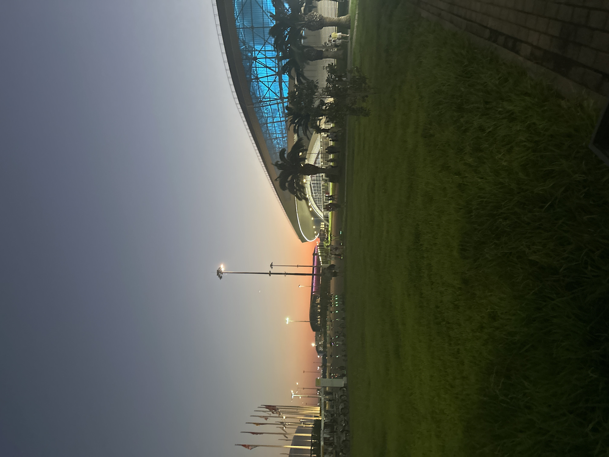 An outdoor, low-angle, full shot captures the Olympic Park in Sochi, Russia, at dusk. The sky transitions from a deep gray at the top to a vibrant orange and pink horizon near the bottom. A large, modern stadium with a translucent blue facade dominates the right side of the frame, its lights creating an ethereal glow. Palm trees stand in front of the stadium, their silhouettes dark against the illuminated structure. The foreground features a well-maintained green lawn, with a paved walkway visible on the far right. To the left, a row of flagpoles stands tall, some bearing national flags. In the mid-ground, a line of electric scooters is parked, suggesting a public transportation or rental service within the park. Several people are visible, walking in small groups or individually, enjoying the evening ambiance. The overall scene conveys a sense of calm and post-event activity in a significant architectural and recreational space.