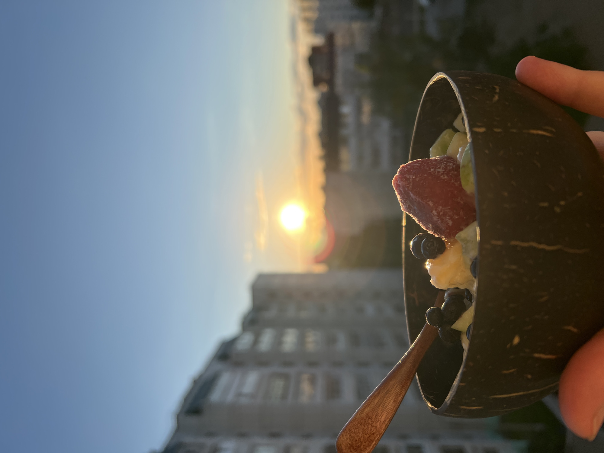 This image, captured during either sunrise or sunset, presents a serene outdoor scene over a city, likely in Chaikovsky, Russia. The composition is horizontally oriented, with the sky dominating the left half and a person's hand holding a food item on the right.
A hand is visible on the right, firmly gripping a dark, textured bowl, which appears to be crafted from a coconut shell. Inside the bowl is a colorful and fresh meal, likely a fruit salad or smoothie bowl. Prominently featured are a large, vibrant piece of red fruit, possibly a strawberry, along with several dark blueberries and lighter-colored fruit chunks, suggesting a healthy snack. A slender wooden spoon is partially visible, resting within the bowl.
The background is an expansive, clear sky, transitioning from a light blue at the top left to a warm, golden hue towards the center where the sun is low on the horizon. The sun is a brilliant, glowing orb, causing a noticeable lens flare with a rainbow effect. Below the horizon line, the blurred silhouettes of several multi-story buildings form a cityscape, indicating an urban setting. The overall lighting suggests the soft, warm glow characteristic of either early morning or late evening. The scene conveys a sense of peaceful enjoyment, perhaps a meal savored with a picturesque view. No text is visible in the image.