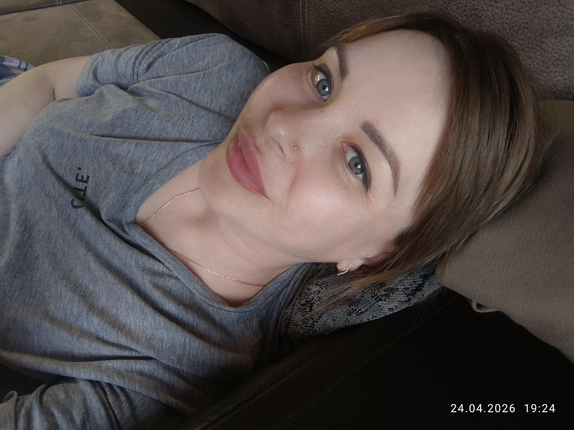 A woman is seen reclining on a dark brown sofa, looking directly at the camera with a gentle, relaxed expression and a slight smile. She has light skin, blue-green eyes, and medium brown hair. A small gold stud pierces her nose, and a delicate gold-colored chain is visible around her neck, along with a small hoop earring in her right ear. She is wearing a grey t-shirt, which has the partial text "CALE" printed on the front. Her eyebrows are well-defined, and she has eyeliner and pinkish-red lipstick. A patterned fabric cushion or throw is visible behind her head. The scene appears to be an indoor setting, likely a home in Chaikovsky, Russia. The bottom right corner of the image includes a timestamp: "24.04.2026 19:24".