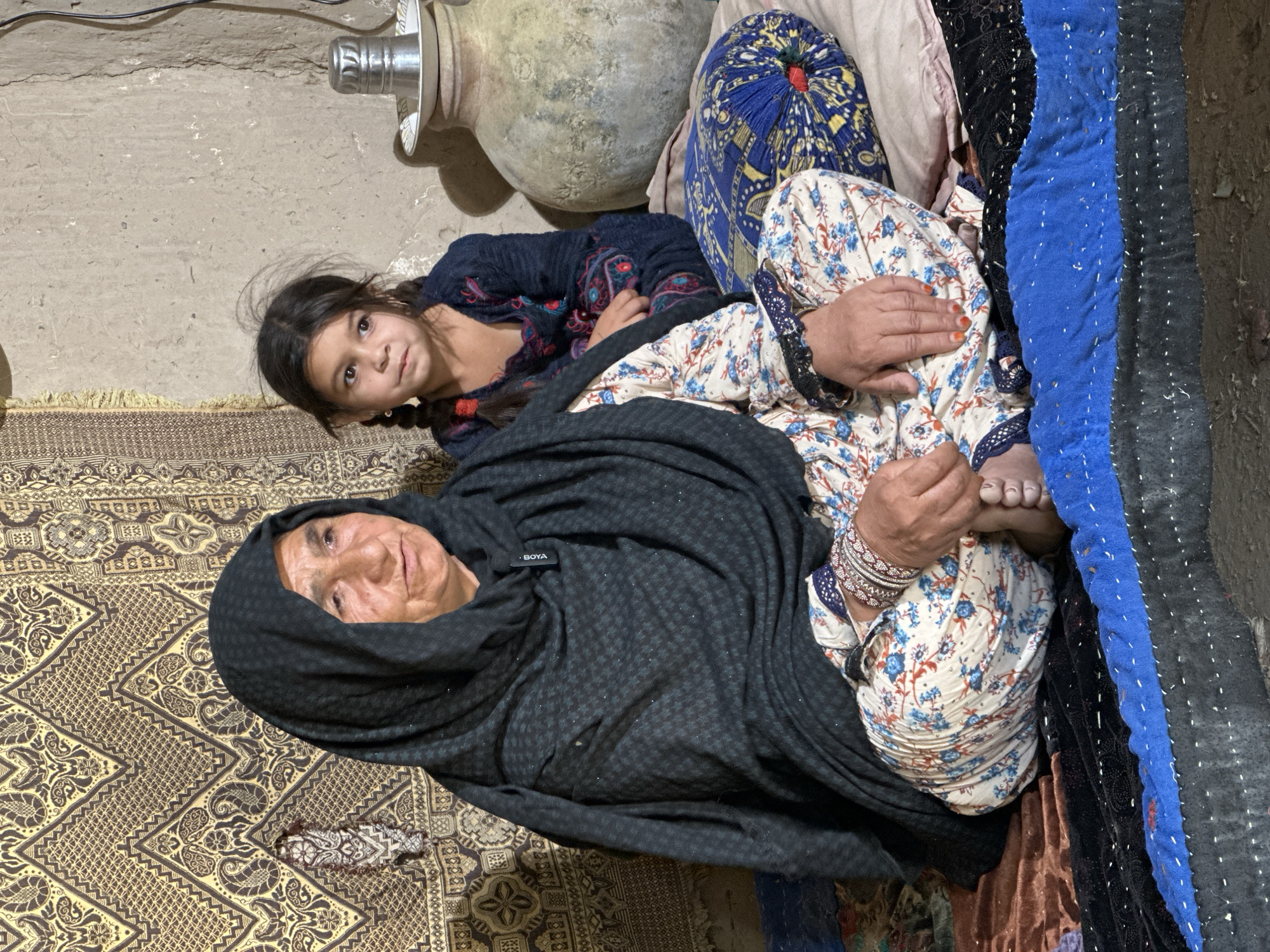 The image shows an elderly woman wearing a black headscarf and a patterned top, lying on an ornate rug, with a young girl with braided hair beside her. The girl is wearing a dark top with embroidery. Behind the girl is a large, round earthenware pot. The woman is resting on pillows and blankets, one with blue patterns. The setting appears to be an interior space. The scene is situated in the city of Quetta City Tehsil, Pakistan.