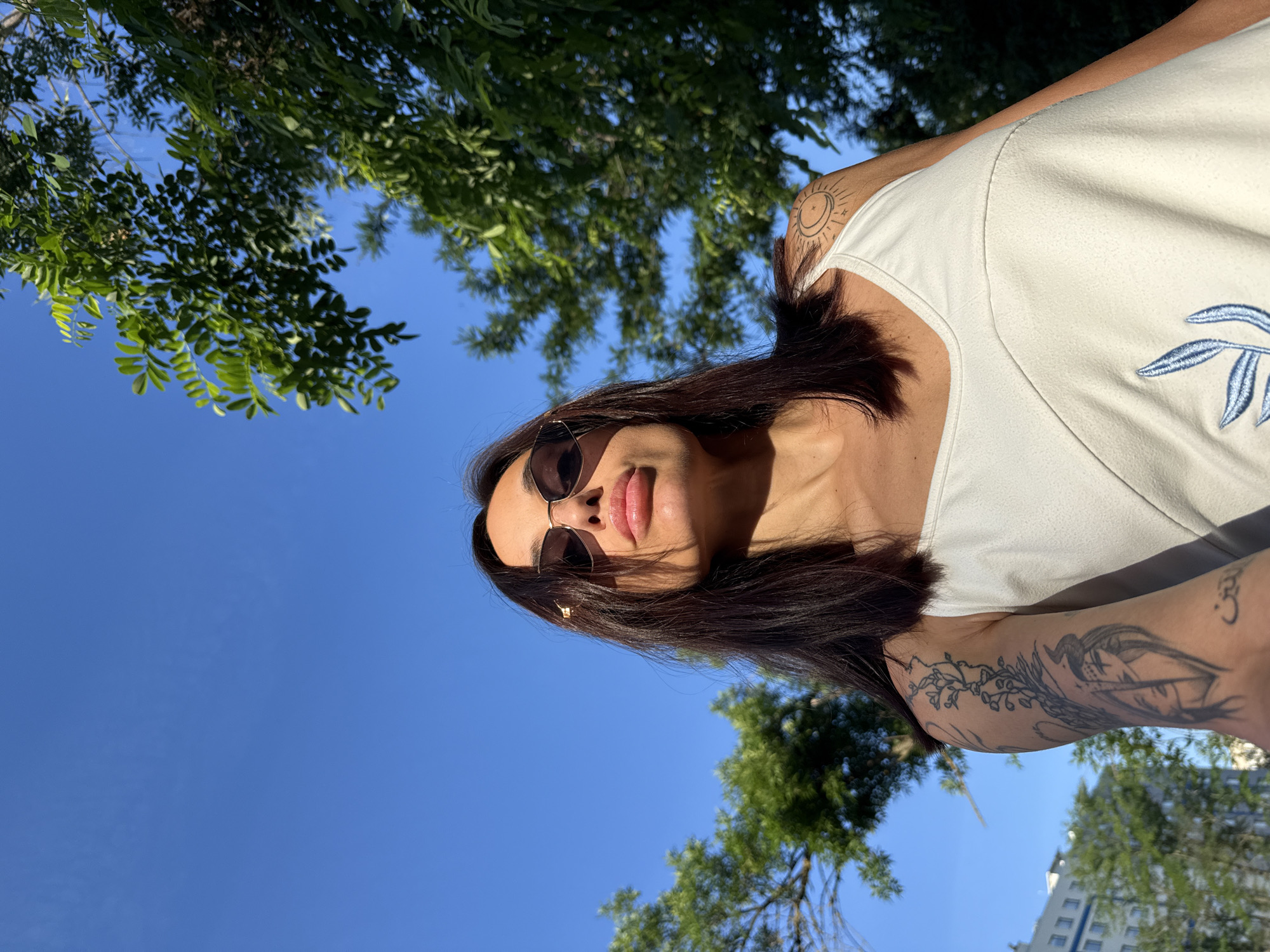 An individual is taking a selfie outdoors on a sunny day. The person is wearing sunglasses and a white camisole with a blue floral design. There are tattoos visible on their arms. The background includes green trees and a clear blue sky, with a building visible in the distance to the left. The image was taken in the city of Pleven, Bulgaria.