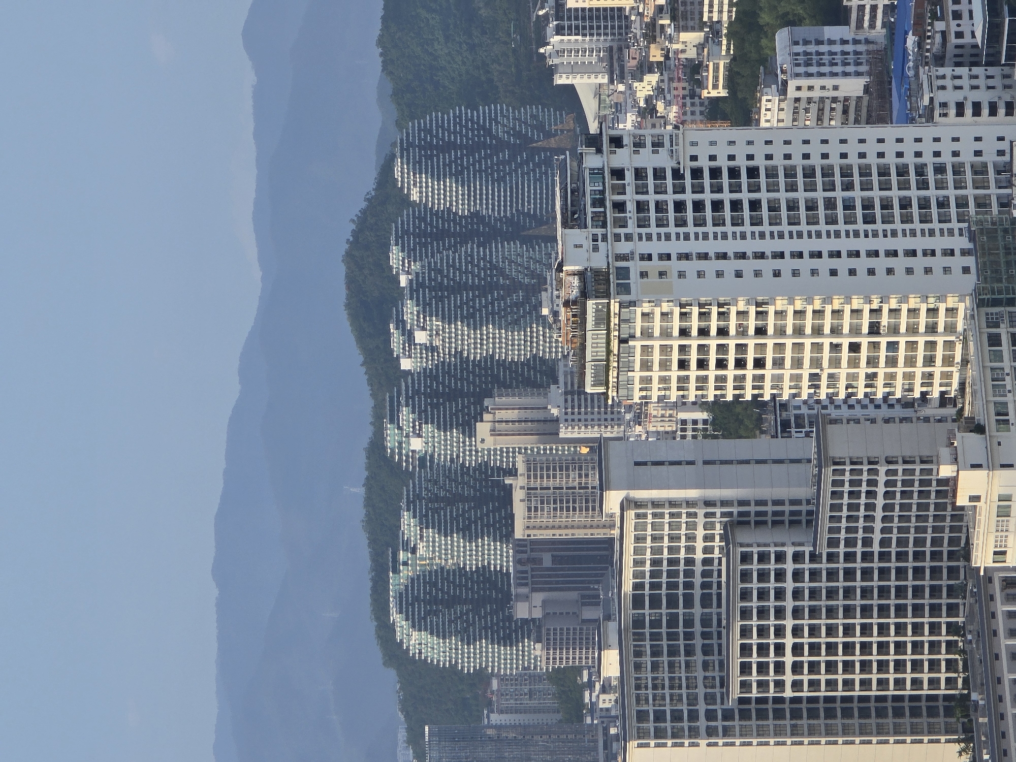 This is a daylight, eye-level, wide-angle, outdoor shot of the architectural landscape of Jiyang District, China, on a clear day. The background features layered, hazy blue mountains, with the closest range covered in lush green trees. In the mid-ground, several unique, tiered buildings with repeating patterns of horizontal white balconies and dark windows are nestled against the mountainside. These structures have a distinctive, undulating, layered appearance. In the foreground, numerous modern, high-rise apartment buildings dominate the scene. These buildings are primarily white or light-colored, with repetitive window grids and various architectural styles, including some with prominent balconies and others with more uniform facades. The sky is a pale, clear blue. No people or vehicles are visible.