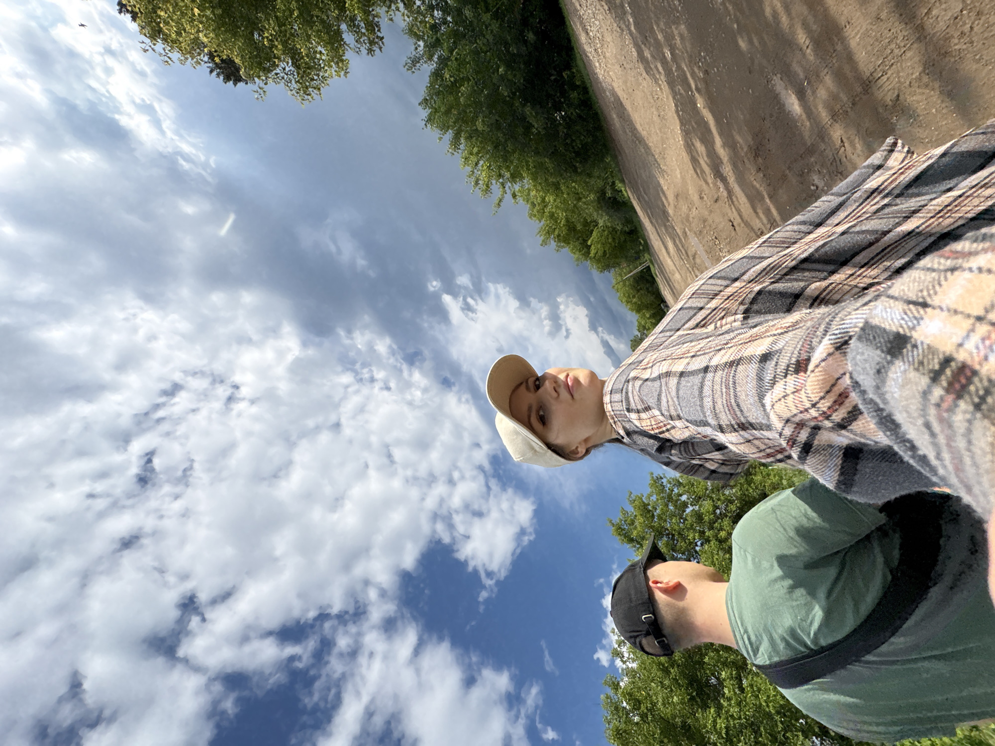 Two individuals, a man and a woman, are captured from a low-angle perspective outdoors. The man, from the back, wears a green t-shirt and a black baseball cap, with a black strap across his shoulder. The woman, facing the camera at an angle, is wearing a light-colored baseball cap and a plaid shirt. Her gaze is directed towards the viewer, and she has a slight smile. They appear to be in an outdoor setting with a dirt path leading uphill, flanked by green trees on either side. The sky above is dramatic, with a mix of bright blue patches and dark, heavy clouds, suggesting a dynamic weather front. The scene suggests a casual outing or walk in a rural or semi-rural area, possibly on the outskirts of the city of Kumyony, Russia, given the presence of natural landscapes like trees and unpaved roads.