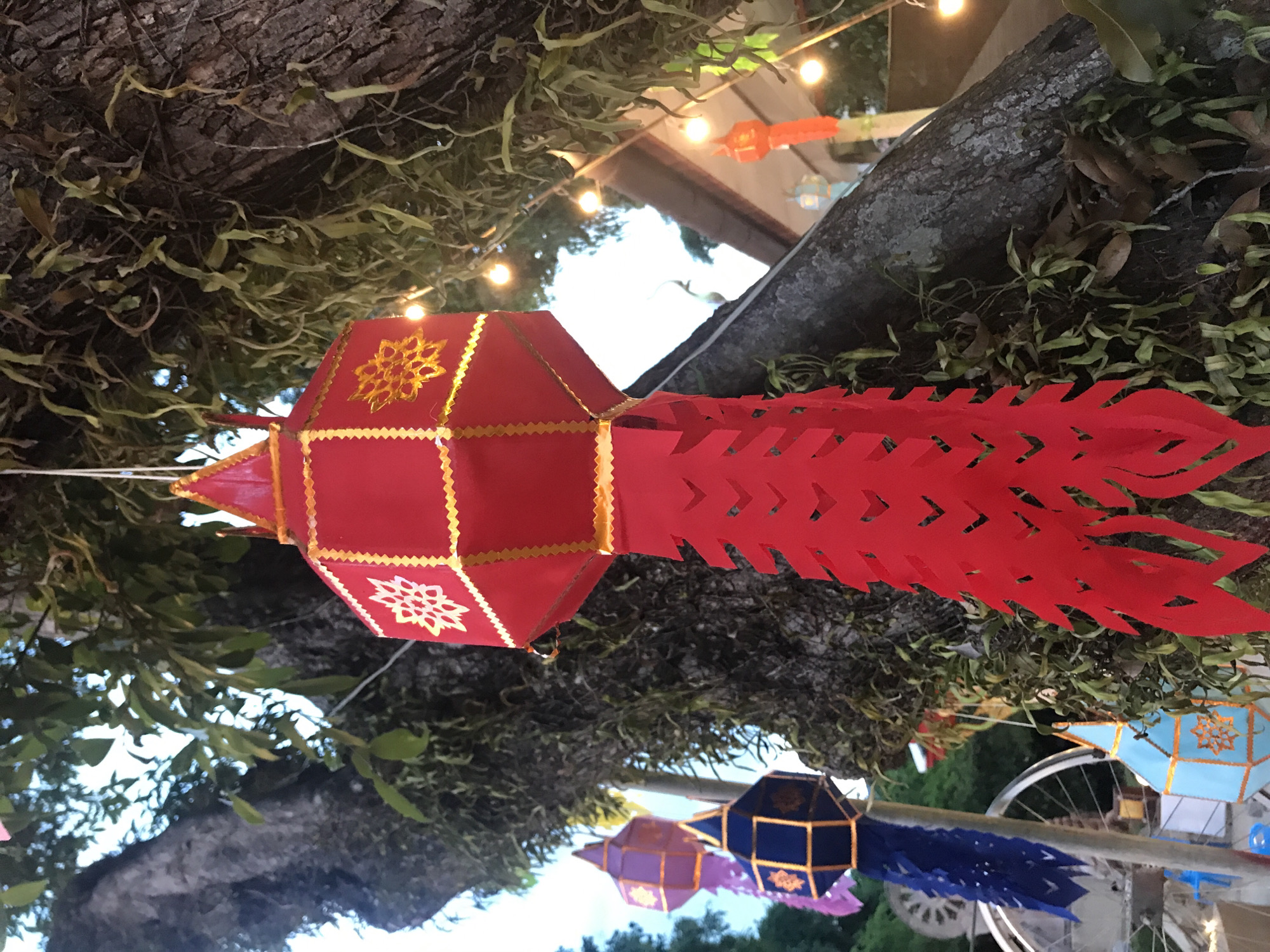 A vibrant scene unfolds in Baan Mae Nam, Thailand, where colorful lanterns adorn a tree. The primary focus is a large, red lantern with intricate gold designs and a long, fringed tail. This lantern hangs suspended by a string, its brightness contrasting against the tree's dark bark and green foliage. In the background, more lanterns of various colors like blue and purple, hang along with a string of lights that adds a festive ambiance to the scene. Parts of bicycles are visible near the lanterns, possibly used for transportation or decoration.