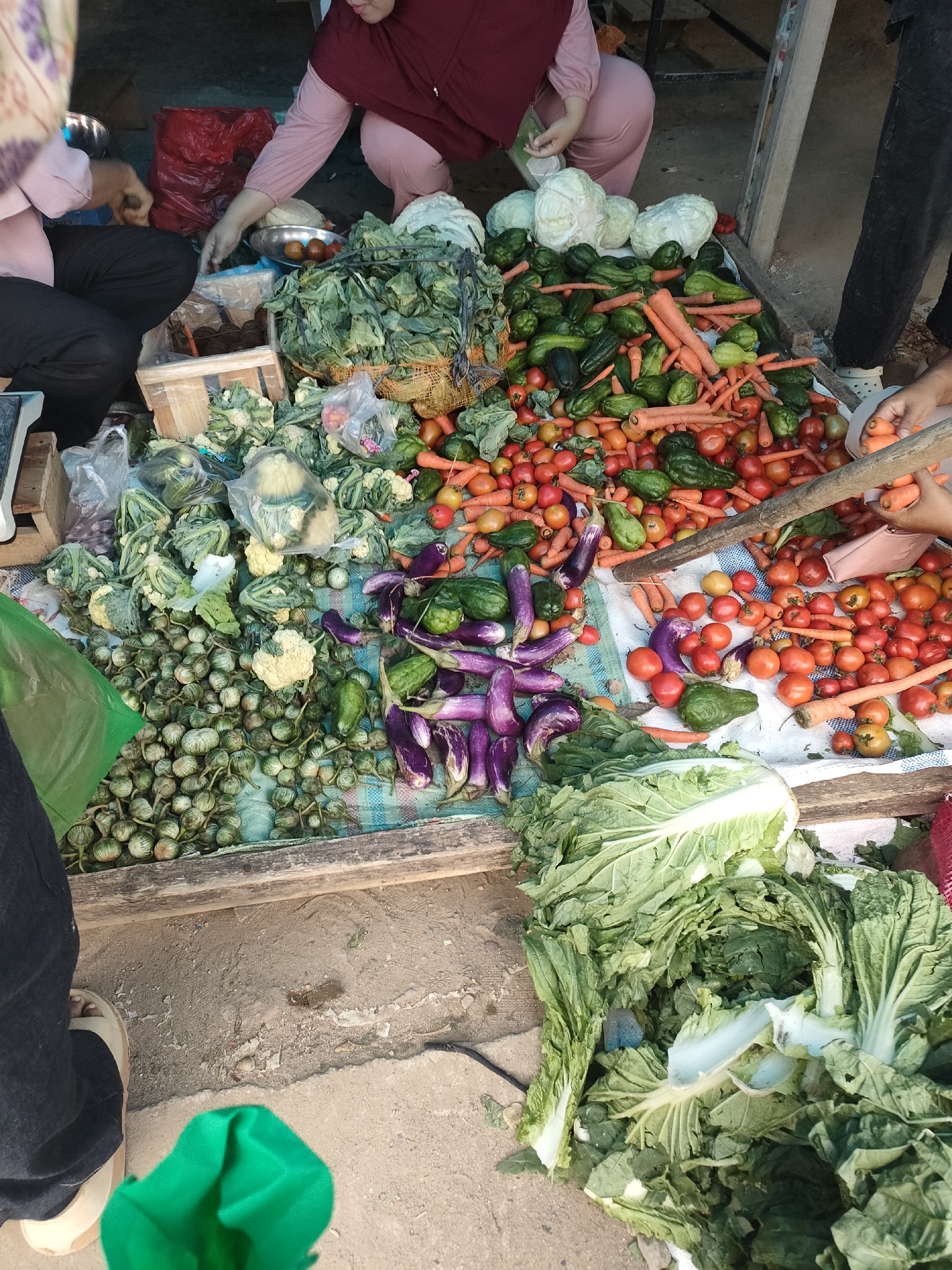The image shows a street-side vegetable market in Sukoyoso, Indonesia. A person in a maroon hijab and pink clothing is crouching to select vegetables. Another person, partially visible on the left, wearing a light-colored top and dark pants, is also engaged with the produce. The market stall is laden with a variety of fresh vegetables including tomatoes, carrots, eggplants, cabbages, and possibly chilies and gourds. The vegetables are displayed on a flat surface, likely a table or a makeshift stall, covered with a patterned cloth. The ground appears to be dirt or a rough pavement. In the background, a red plastic bag and other market elements are visible, suggesting a bustling marketplace environment.