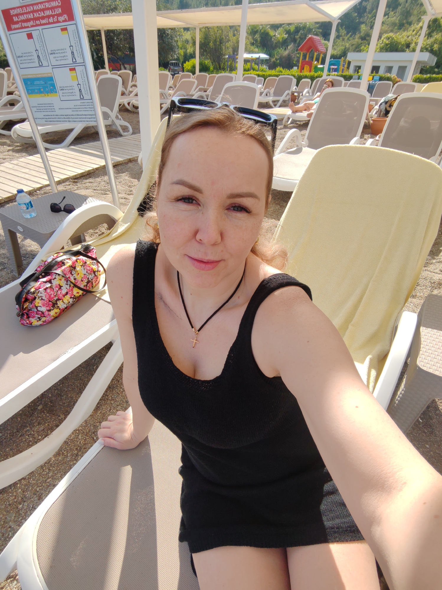 A woman takes a selfie on a lounge chair at a beach in Alanya, Turkey. She has light skin, reddish-blonde hair pulled back, and is wearing a black tank top dress and a necklace with a cross pendant. Her sunglasses are perched on her head. In the background are numerous other lounge chairs under canopies, some occupied by people, and a playground. A small table next to the lounge chair holds a water bottle and a pair of sunglasses, with a floral patterned bag on the lounge chair. The beach has light colored sand and is well-maintained.