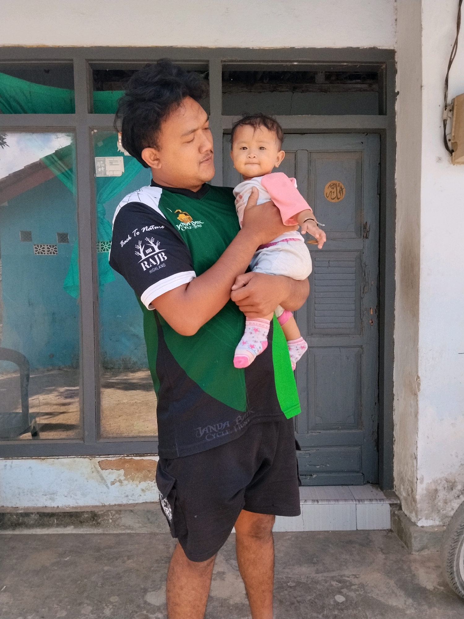 A man stands holding a baby in front of a weathered blue door in Panggungan, Indonesia. The man is wearing a green and black t-shirt and black shorts. The baby is wearing a pink and white outfit. The background features a building with a blue wall and windows, suggesting an outdoor or semi-outdoor setting. The overall scene appears to be a candid moment captured outside a residence.