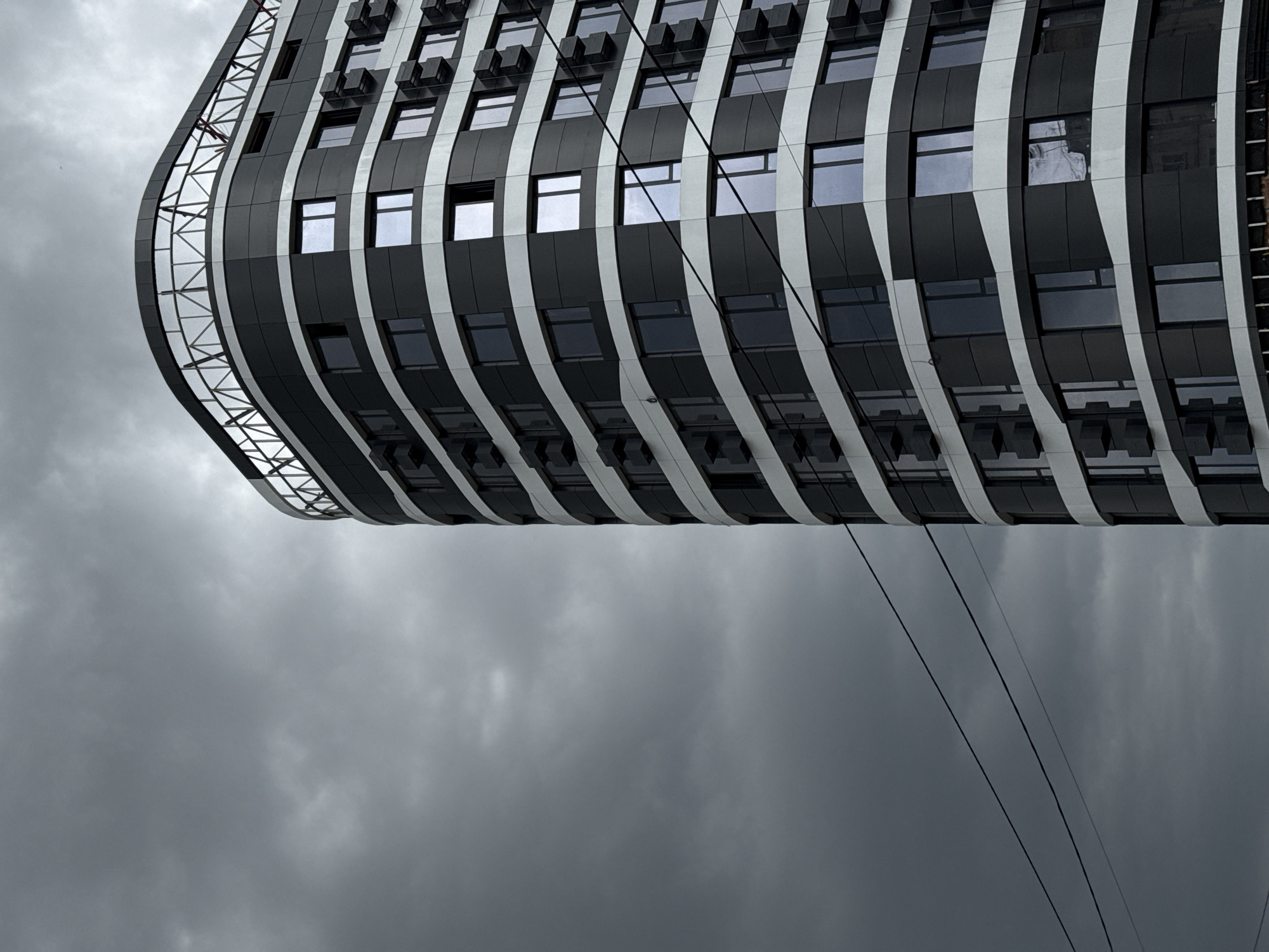 A low-angle, full shot captures the facade of a modern, curved building against a dark, overcast sky. The building's exterior is a striking pattern of black and white horizontal bands, with rows of dark windows interspersed. Cubical air conditioning units are visible protruding from the facade on many floors. Two dark electrical wires run diagonally across the frame in the lower left portion, cutting across the sky and the building. The upper edge of the building features a distinctive open-air structure with a metallic, lattice-like railing. The overall mood is somber due to the heavy cloud cover.