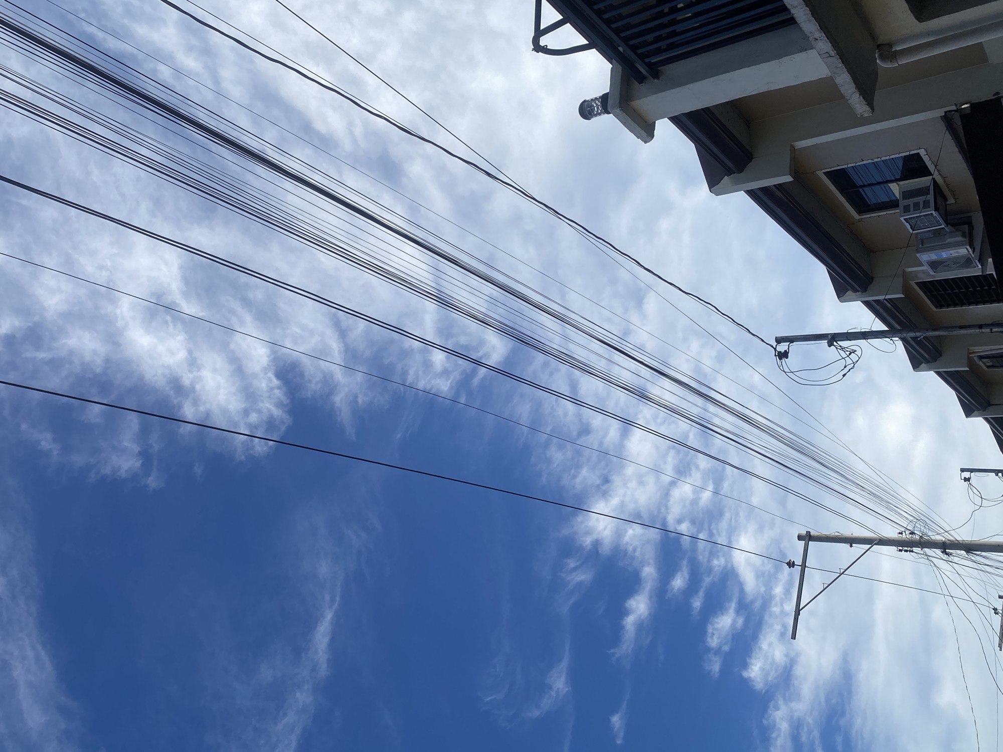 The image captures a street scene in Davao City, Philippines, dominated by a bright blue sky with scattered wispy white clouds. Overhead, a tangle of black electrical wires stretches across the frame, running parallel and diagonally. To the right, the upper floors of beige residential buildings are visible, featuring dark-tiled roofs and windows. Some of the buildings have air conditioning units installed. A black metal fence and a decorative lamppost are also present on the right side of the image. On the left, a utility pole with multiple wires connected to it stands against the sky. The overall impression is of a typical urban environment under a partly cloudy sky.