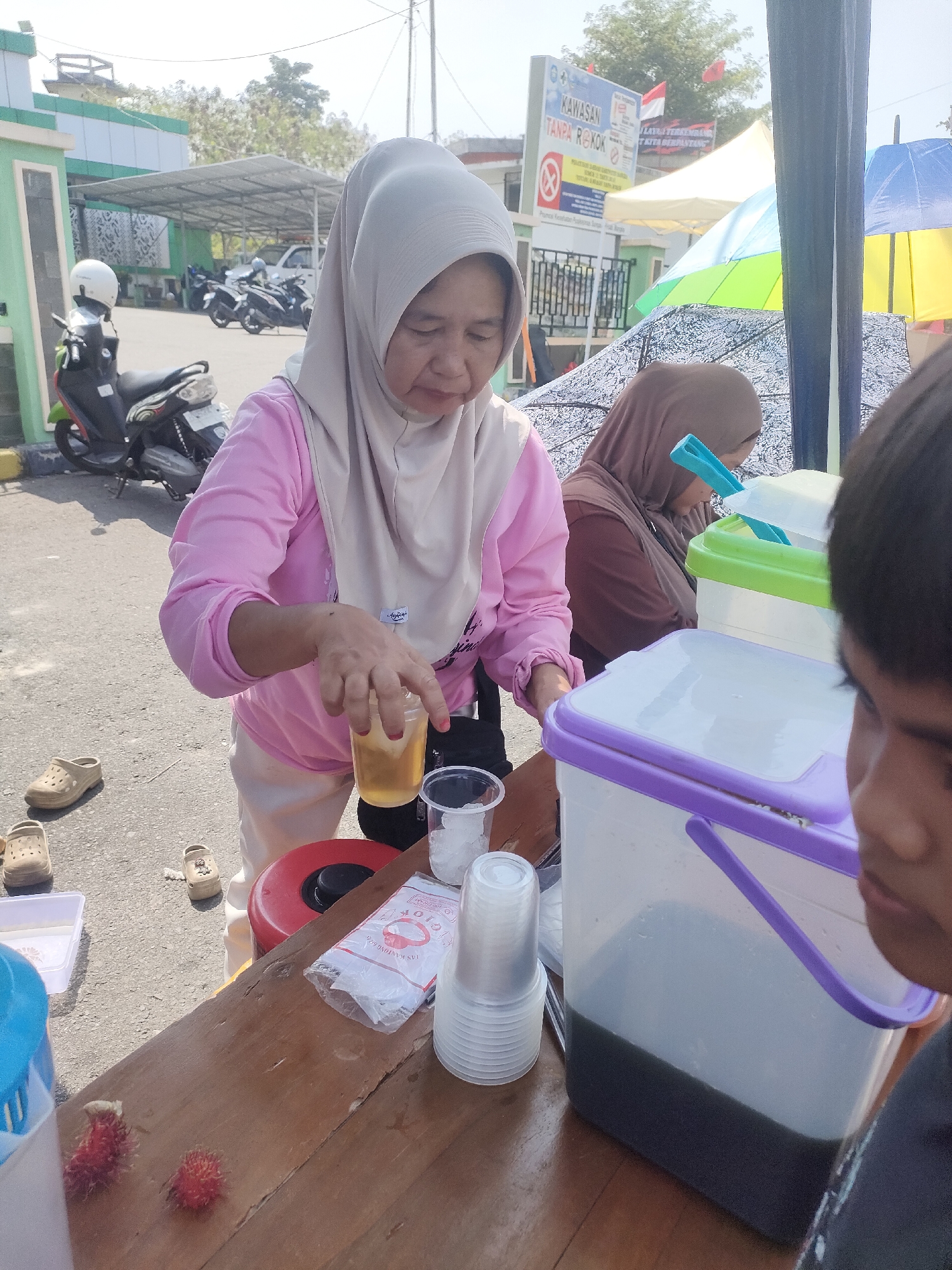 In Sungailiat, Indonesia, a vendor with a light brown hijab and a pink shirt serves drinks from a wooden table outdoors. She is pouring liquid from a glass into a plastic cup. To her left, a person wearing a darker hijab is visible behind the table. In front of the table, several plastic cups, a bag of plastic bags, and two rambutans are arranged. A large transparent cooler with a purple lid is to the right of the table, containing a dark liquid. In the background, motorcycles are parked under a canopy, and a sign with Indonesian text is visible. The scene appears to be a street-side stall on a sunny day.