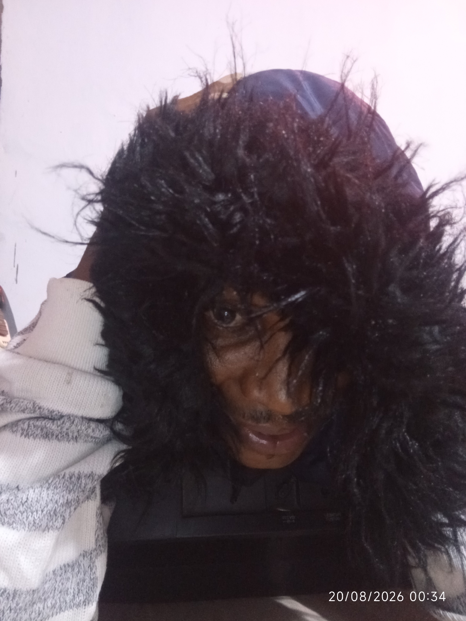 A close-up, low-angle shot captures a person's face from the shoulders up, with the individual looking towards the camera. The person is wearing a thick, black, fur-lined hood that partially obscures their face, with strands of the fur falling forward. Their right eye is visible, along with a portion of their nose and mouth, which appears to be slightly open. The person is also wearing a white and grey striped sweater or hoodie. The background is a plain, light-colored wall. A timestamp in the bottom right corner reads "20/08/2026 00:34". The scene is indoors, likely a residential setting in Adankolo, Nigeria.