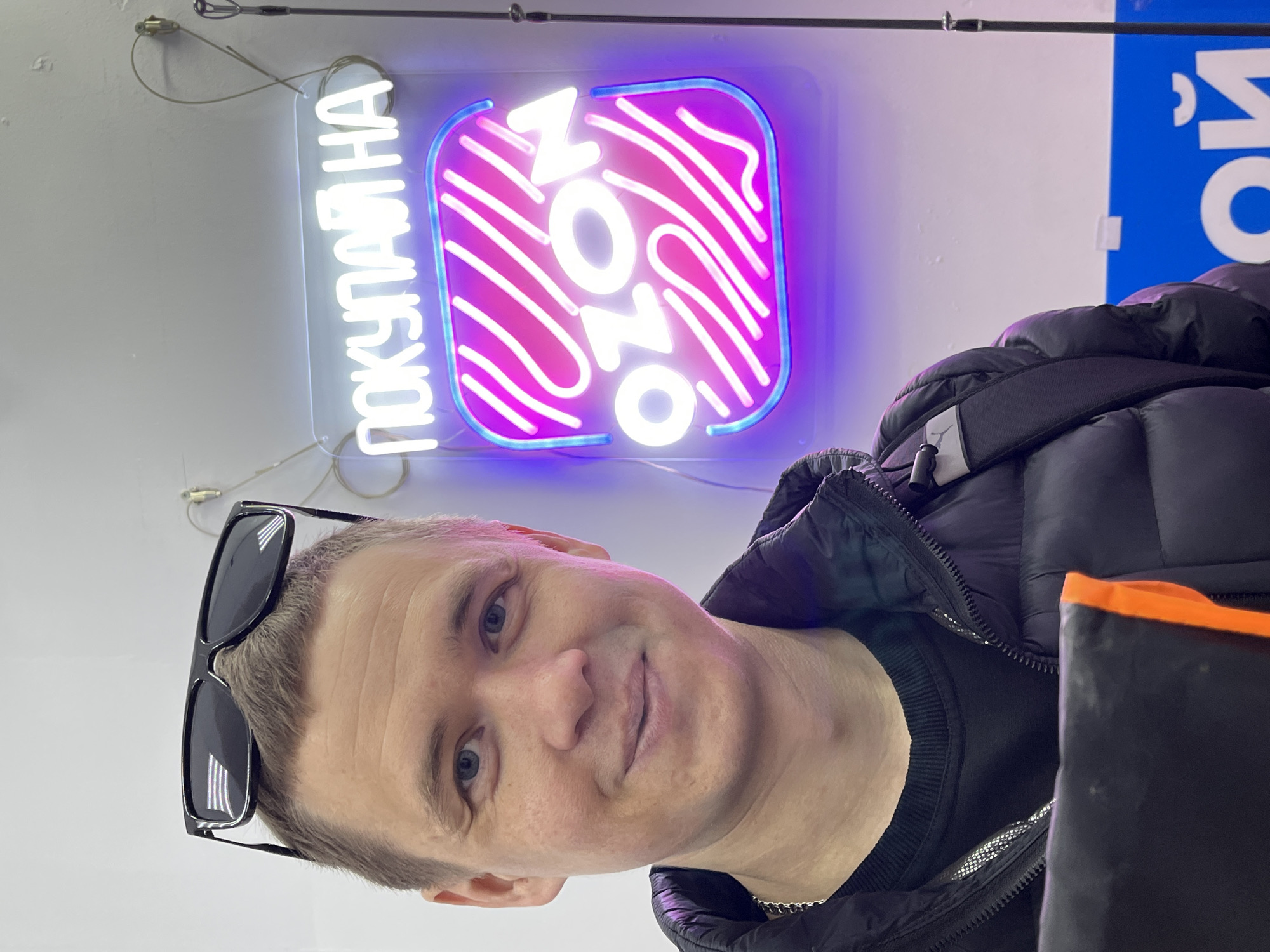 A man with short blond hair and blue eyes poses for a selfie indoors. He is wearing a black puffer jacket, a black t-shirt, and sunglasses resting on his head. In the background is a neon sign for "Ozon" in Russian and a partially visible blue sign. The photo appears to be taken in a commercial space, possibly a store or office. While there is no specific location information, the sign suggests this is somewhere in Russia. Due to the absence of distinguishing geographical markers, it isn't possible to identify the exact location in Chaikovsky, Russia.