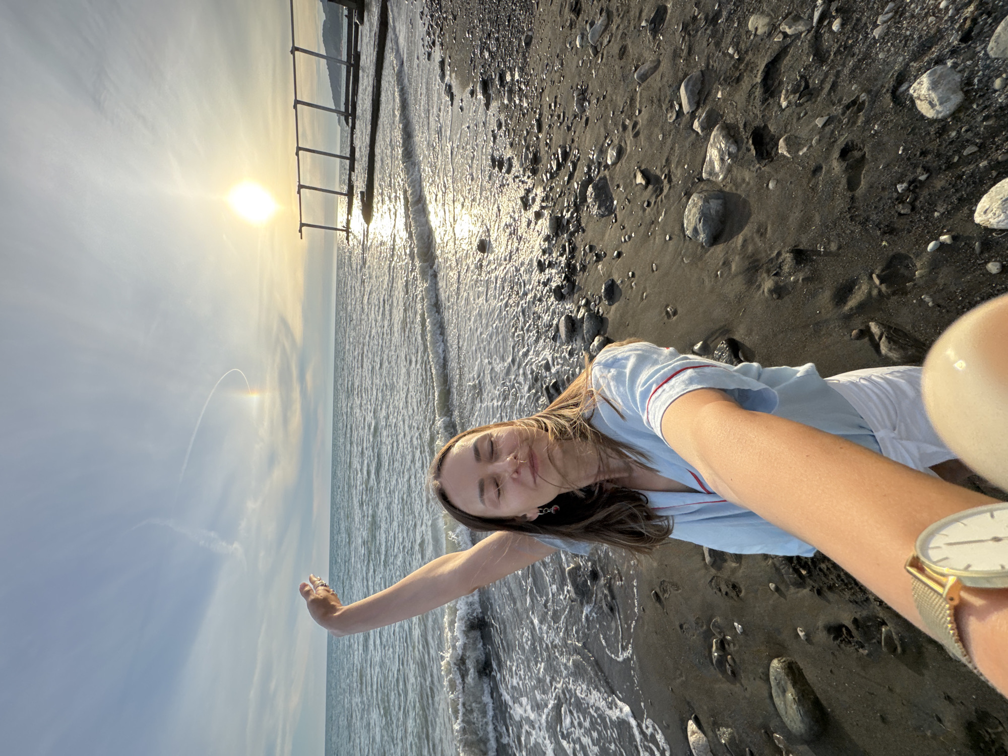 A woman is taking a selfie on a pebble beach in Sochi, Russia, with the Black Sea in the background. The sun is setting, casting a golden glow on the water and the sky, which features a faint rainbow. The woman is wearing a light blue pajama-like top with red trim and white shorts, and a gold watch on her left wrist. She has her eyes closed and her right arm raised, with her hand open. A pier-like structure is visible in the distance on the water. The beach is covered in dark sand and various sized pebbles, with some footprints visible in the sand.
