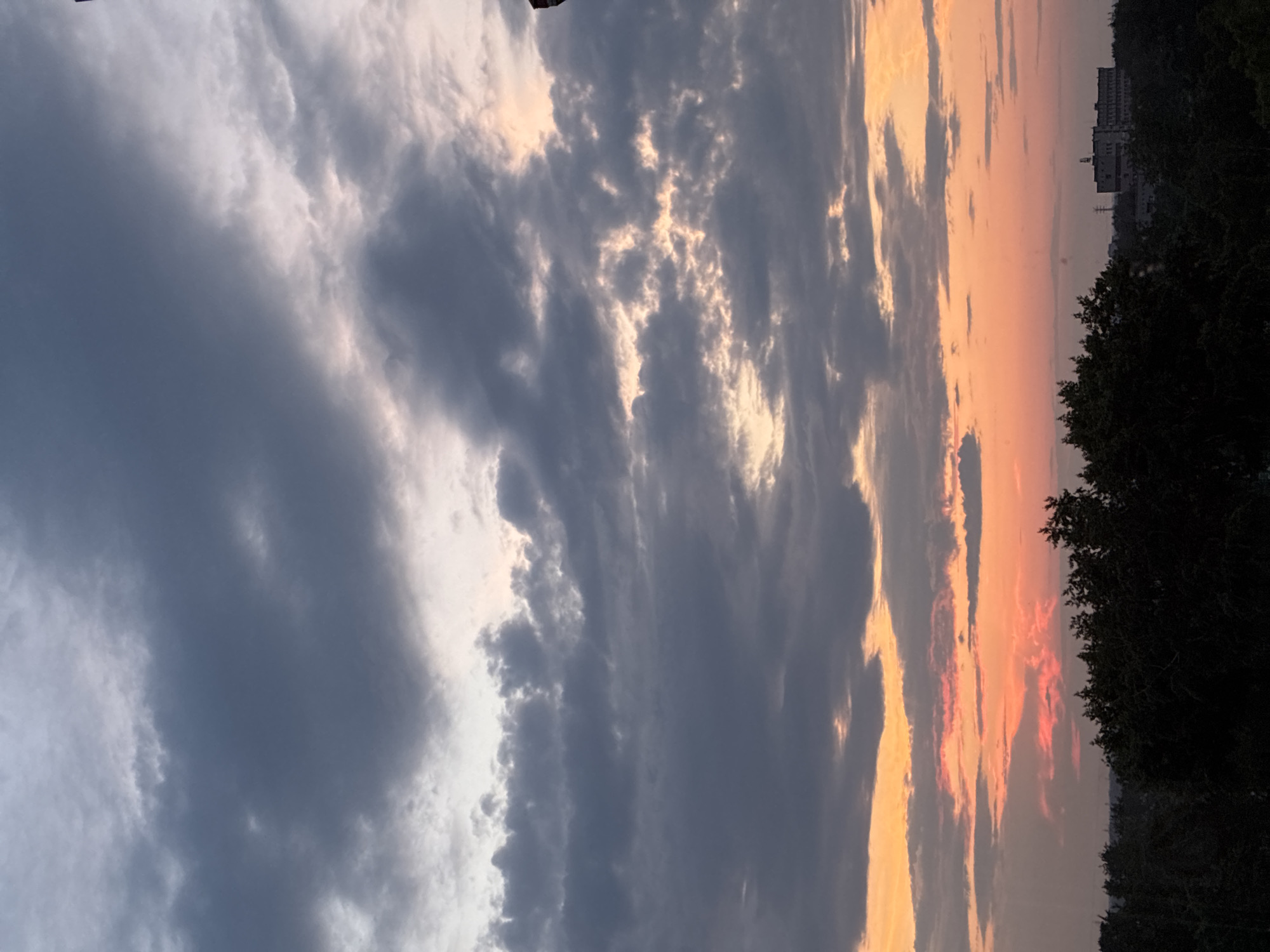 A wide shot captures a dynamic sky filled with a gradient of colors as the sun sets. The upper portion of the frame is dominated by dark, heavy clouds tinged with gray, suggesting an impending storm or the aftermath of rain. As the eye moves downwards, these clouds give way to lighter, more wispy formations, interspersed with streaks of bright orange and pink hues, indicative of the setting sun's rays breaking through.
The horizon line is low in the frame, emphasizing the vastness of the sky. Along this line, a silhouette of dark green trees forms a dense canopy, obscuring the ground below. Beyond the trees, a few distant buildings are visible, including a larger, block-like structure that appears to be a multi-story building or complex, hinting at an urban or suburban environment. A solitary dark vertical object, possibly a utility pole or antenna, stands out against the colorful sky in the far upper right corner of the image. The overall scene is atmospheric, showcasing the dramatic beauty of a twilight sky over a silhouetted landscape.
