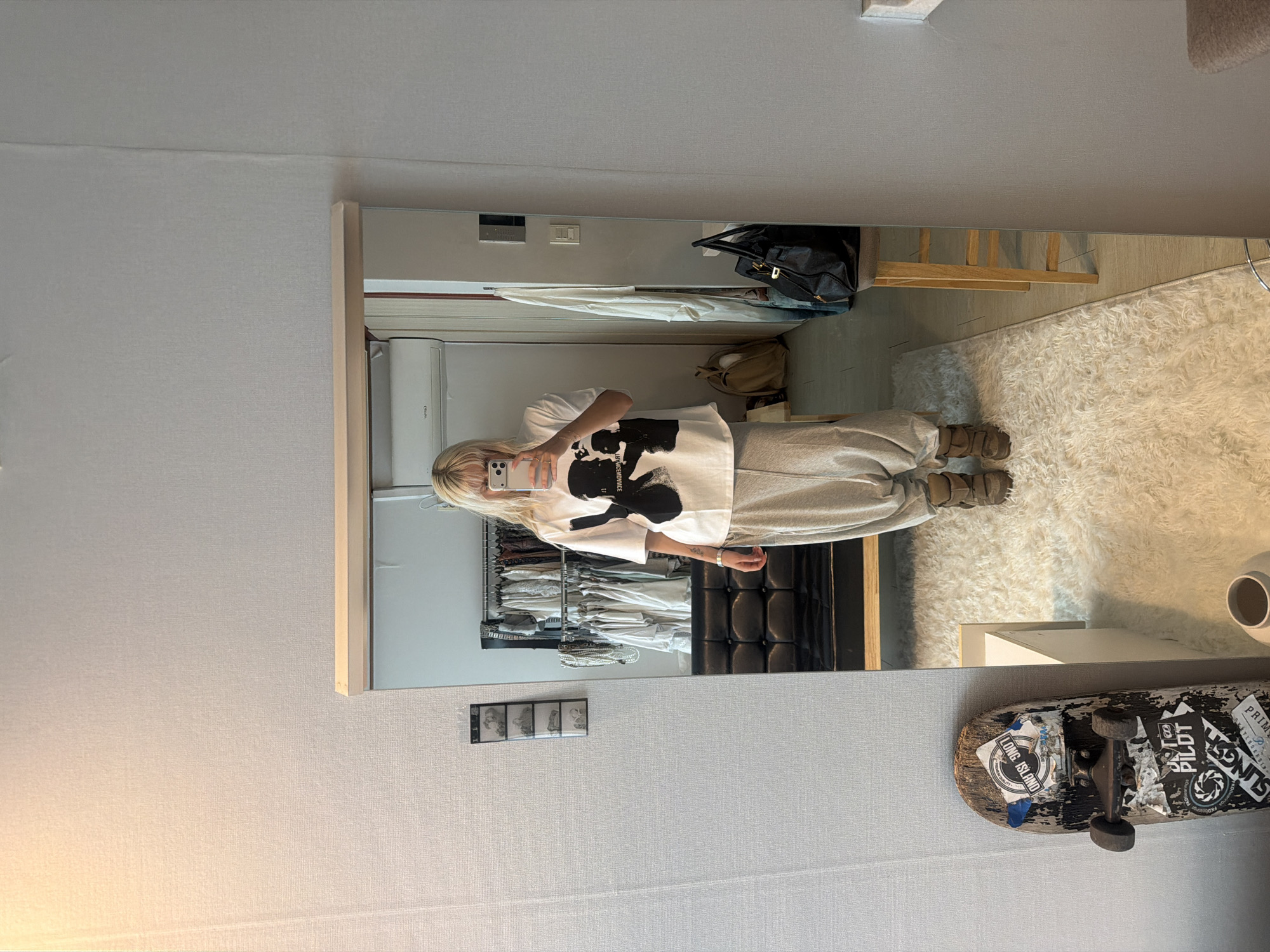 A full-length mirror selfie captured indoors in what appears to be a bedroom or dressing area in Seoul, South Korea. A person with long blonde hair, seen in the mirror's reflection, is wearing an oversized white t-shirt with a black graphic design featuring a silhouette of a person's head and partially obscured text, including "OF THE CENTURY" with an inverted 'M'. They are also wearing light grey sweatpants and chunky brown sandals or clogs, and are holding a light-colored smartphone to take the picture.
The room features light, textured wallpaper. In the mirror's reflection, behind the person, an organized open closet or clothes rack filled with numerous garments is visible. To the right, a black handbag rests on a surface, and a beige shoulder bag is also present. The floor has a white, shaggy rug. Adjacent to the mirror on the actual wall, a skateboard with several stickers (including "LONG ISLAND," "PIT PILOT," and "SLING") leans against the wall next to a white ceramic vase or cup. A strip of four small framed photos is mounted on the wall to the left of the mirror. An air conditioner unit is mounted on the wall in the reflection. The overall scene suggests a casual, everyday moment.