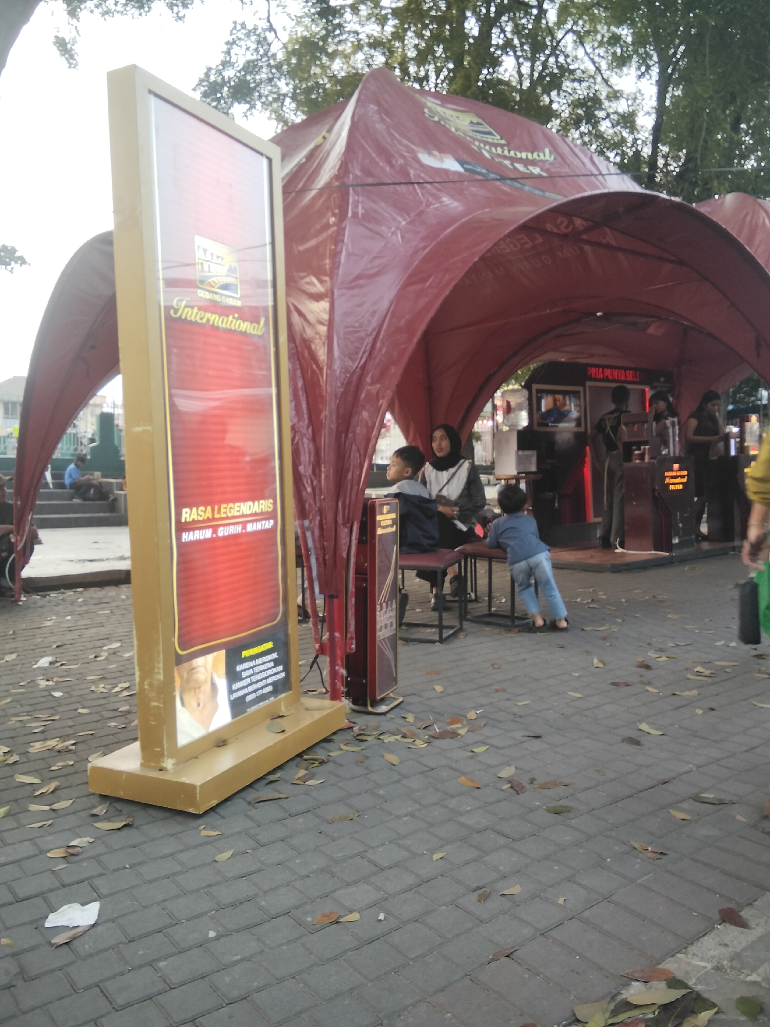 This is a medium-shot, eye-level, outdoor photograph of a temporary promotional tent with a red tarp. The tent is set up on a paved area with fallen leaves. A large, illuminated red and gold sign stands to the left of the tent, displaying text that appears to be "GUDANG GARAM International" and "RASA LEGENDARIS HARUM, GURIH, MANTAP". Below this, there is an image of an older man and warning text.
Inside the tent, several people are visible. A woman in a white headscarf and black jacket is seated on a stool, facing left. Two young boys are near her; one is sitting on her lap, and the other, wearing a blue shirt and jeans, is standing and leaning towards the tent's counter. Behind them, a dark-haired woman in a black top is standing behind a counter, and another person, possibly male, is standing further back and to the right, wearing a dark shirt and pants. A television screen is mounted on the back wall of the tent, displaying an image.
The environment suggests an outdoor public space, likely a street or a park, with trees visible in the background and dappled sunlight. The presence of the "Gudang Garam International" branding indicates this is likely a promotional event for cigarettes, a common sight at public venues in Indonesia. The text "PRA PONYA SELE" is also visible on the tent's interior wall. Given the context of Gudang Garam, a prominent Indonesian tobacco company, this scene could be a promotional event in a public area within the city of Cisaat, Indonesia, known for its association with Gudang Garam's manufacturing facilities.