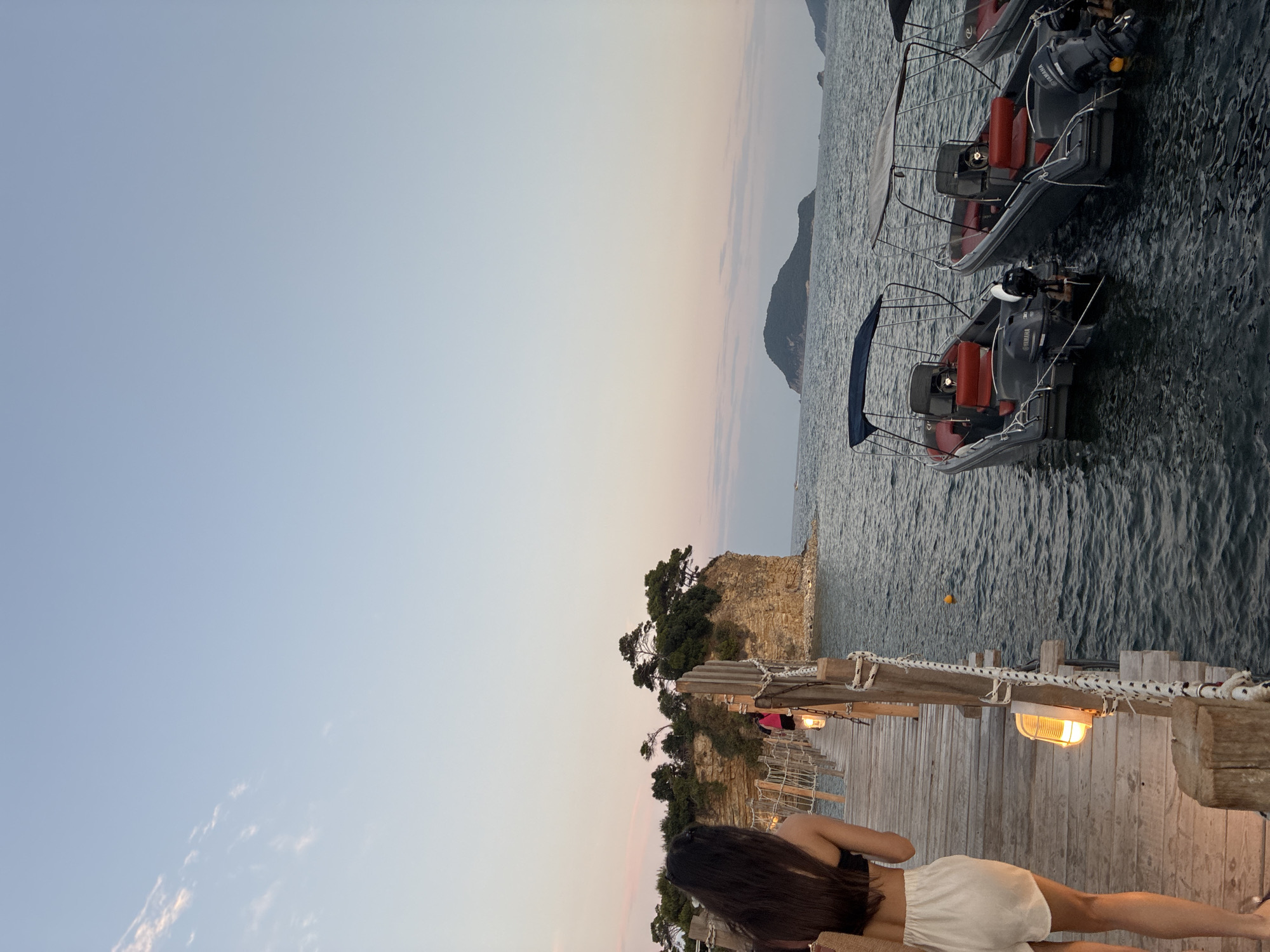 The image depicts a wooden pier extending into the sea, with a woman in a black bikini top and white shorts walking away from the camera on the left. Several boats are docked in the water to the right, and a rocky island with trees is visible in the background. The sky is clear and a soft pink and orange hue suggests either sunrise or sunset. The location appears to be a coastal area, possibly a tourist destination. Based on the visual cues, it is likely in Laganas Municipal Unit, Greece, known for its scenic beaches and islands.