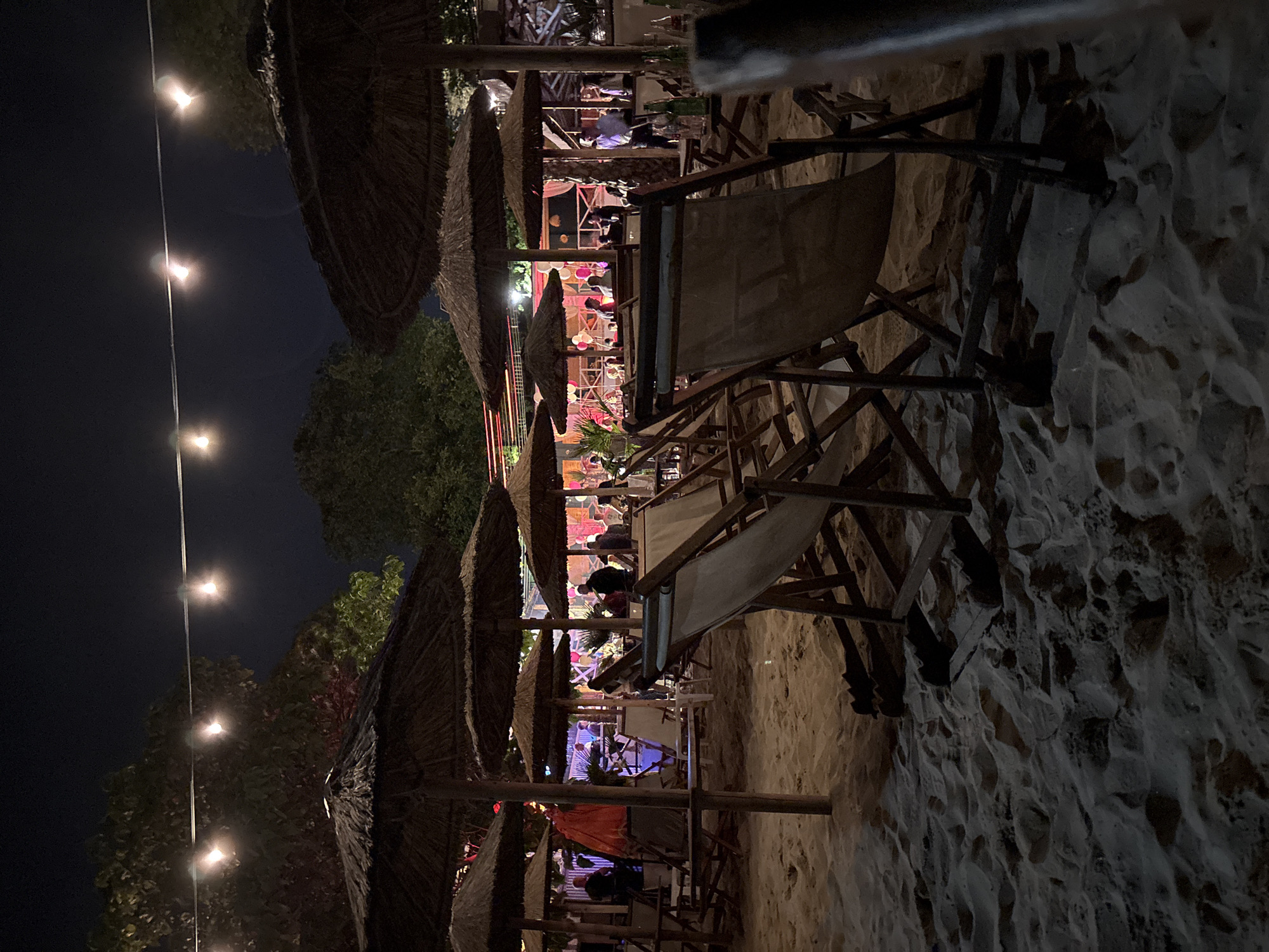 The image depicts a nighttime outdoor scene in Varna, Bulgaria, at a beach-themed establishment. The ground is composed of sand, showing numerous footprints and undulations. In the foreground and midground, multiple dark-thatched umbrellas with wooden poles are prominent, casting shadows. Numerous wooden lounge chairs with canvas slings are arranged on the sand, some appearing empty, with a particularly clear view of one chair's frame on the right side. Overhead, a string of warm-toned incandescent bulbs extends horizontally across the frame, providing ambient illumination.
In the midground, beyond the umbrellas and chairs, various individuals are visible, mostly as silhouettes or partially illuminated figures, indicating a social gathering. Some appear to be standing, others seated. A notable reddish-orange fabric structure or light installation is visible behind one of the central umbrellas. In the background, an elevated structure, likely a bar or stage area, is brightly lit with various colored lights, including pinks and whites. This structure features decorative elements such as white and pink balloons and draped fabric. Several people, some appearing to be staff members in light-colored shirts, are present on and around this platform. Dark foliage of trees is visible against the night sky in the upper background. Scattered tables with bottles are also discernible in the midground and background.