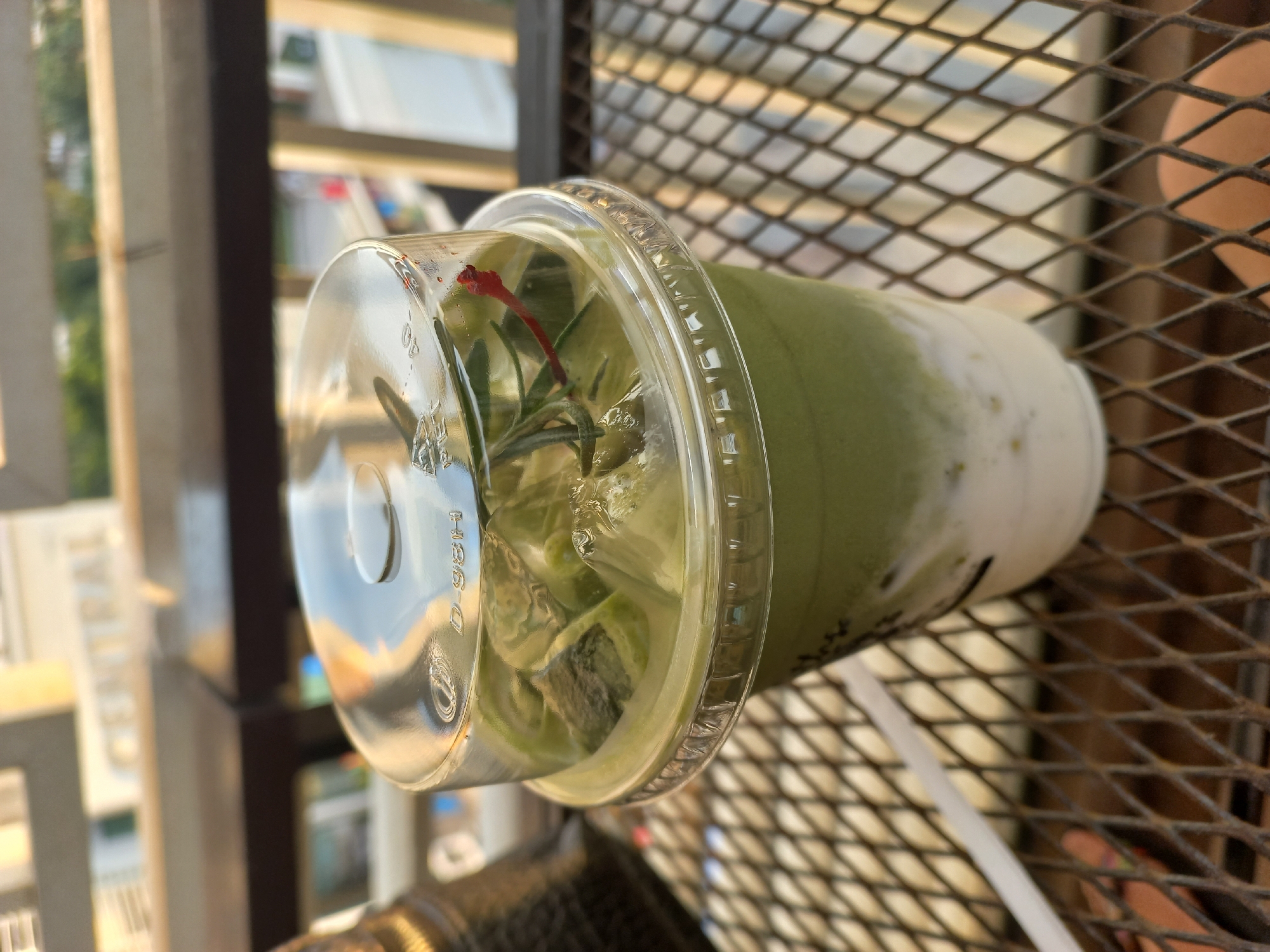 A high-angle, close-up shot shows a plastic cup of iced matcha latte with a clear lid, placed on a metal mesh surface. The cup is filled with a light green liquid, ice cubes, and a small red cherry. Outside, the city of Pattaya City, Thailand, is visible in the background, including a modern high-rise building and parts of the sky.