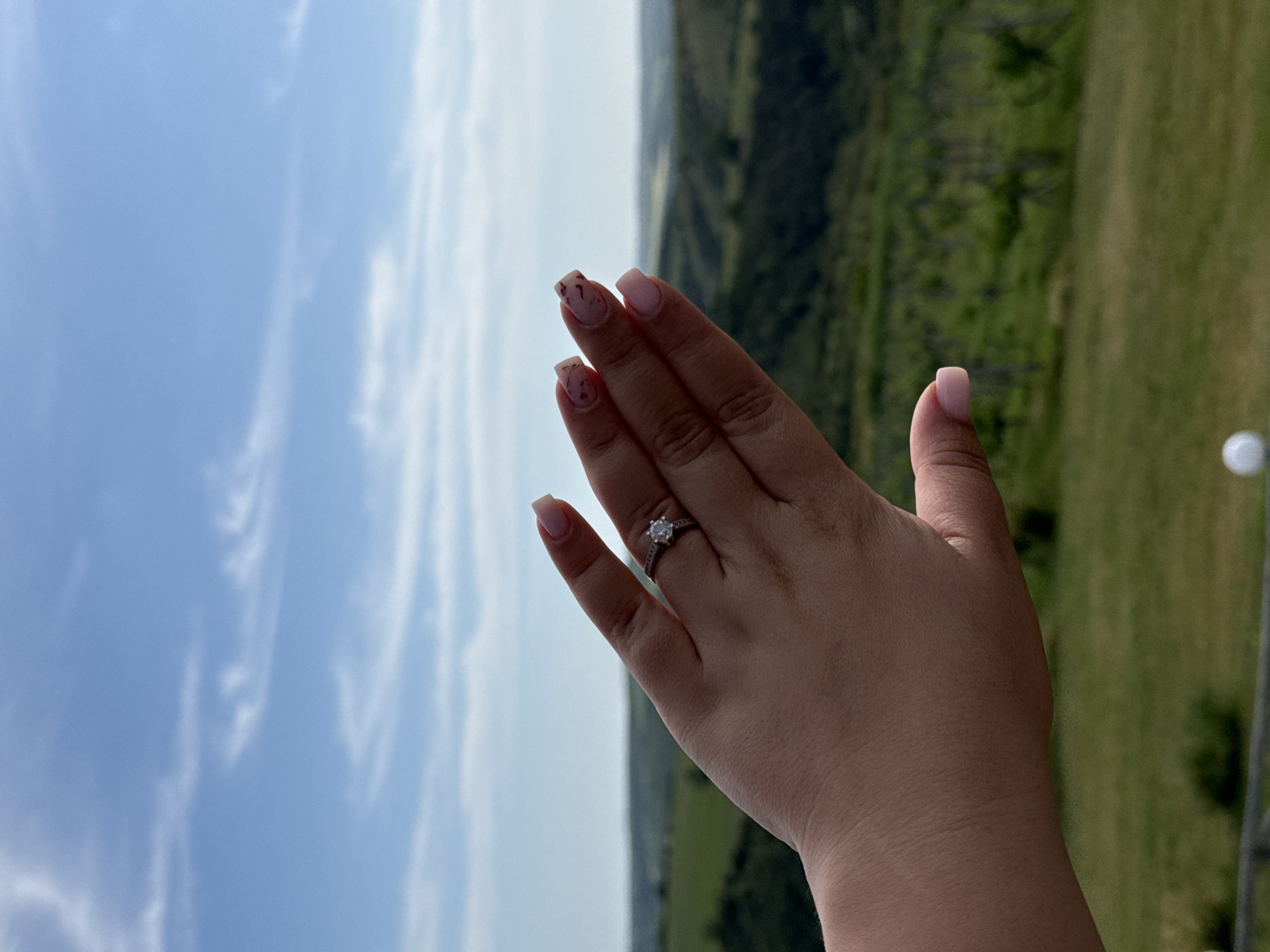 A woman's left hand is held up against a backdrop of sky, trees, and rolling hills, presenting an engagement ring on her ring finger. The ring features a round diamond set in a silver-colored band. The nails on her fingers are painted with a light pink base and a design in a darker color. The location is assumed to be in Mechka, Bulgaria, though this is not directly verifiable from the image itself.