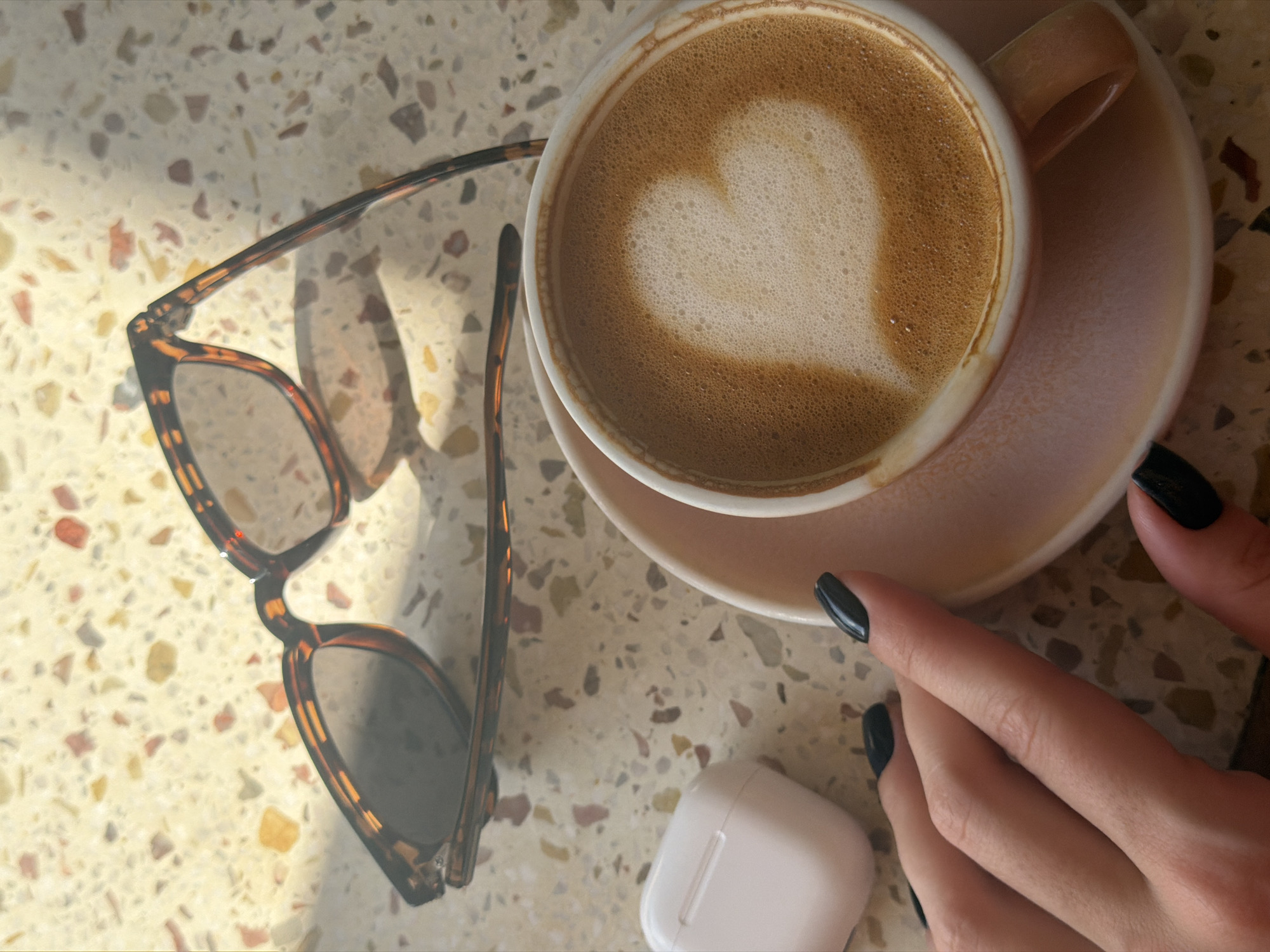 An overhead shot captures a serene tabletop scene on a sunny day. The main subjects include a cup of coffee with latte art, a pair of sunglasses, a person's hand, and a white earbud case, all resting on a light-colored, speckled table.
The coffee cup, a light-colored ceramic, is positioned towards the upper right of the frame, sitting on a matching saucer. The coffee inside features a distinct heart-shaped design in its white foam, suggesting it's a latte or cappuccino. To the right of the cup, a person's hand with dark, glossy black nail polish is visible, fingers gently resting near the saucer.
On the left side of the table, a pair of tortoiseshell-patterned sunglasses with dark lenses is laid open. Below the coffee cup and slightly to the left of the hand, a white, rectangular earbud charging case is present. The table surface is illuminated by strong, dappled sunlight, creating patterns of light and shadow, indicating a bright daytime setting. The overall scene evokes a sense of calm and leisure, perhaps a coffee break outdoors or in a sunlit indoor space. There is no visible text in the image.