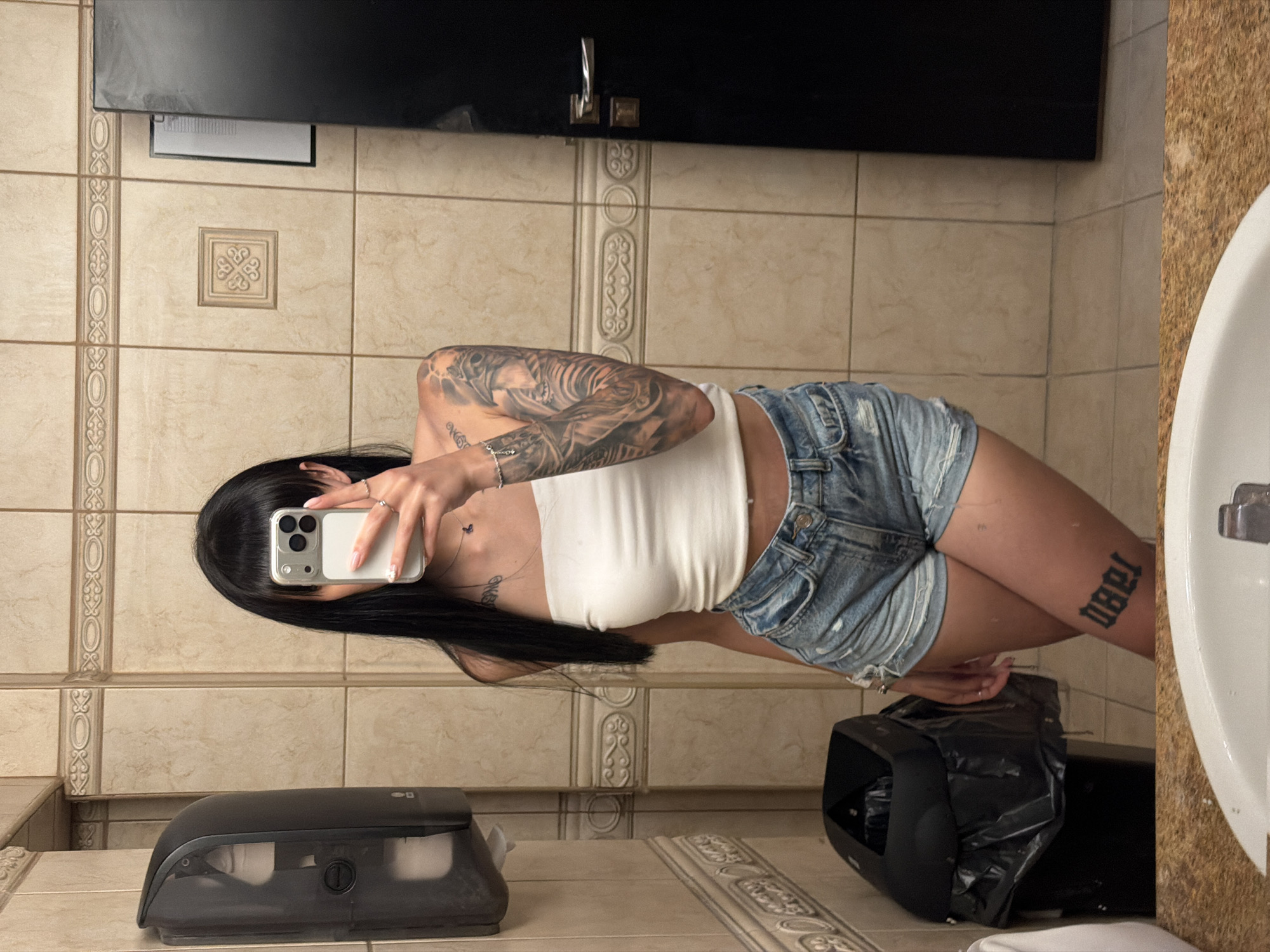 A young woman is captured in a mirror selfie within what appears to be a public restroom in Plovdiv, Bulgaria. Her face is obscured by a silver-colored smartphone with a clear case, held in her left hand. She has long, dark hair with bangs and is wearing a white strapless crop top and light blue distressed denim shorts. Visible tattoos include a full sleeve on her right arm, depicting what appears to be a warrior or skull motif; a small, dark tattoo resembling a butterfly on her upper chest; and a prominent gothic-style script tattoo on her left thigh, which reads "YƎBO" when viewed directly. Her nails are manicured in a light color, and she wears multiple rings on her left hand and a bracelet on her right wrist.
The restroom features light beige square wall tiles with a decorative horizontal border tile and a single ornate accent tile. To the left, a dark gray automatic paper towel dispenser is mounted on the wall, with a black trash can containing a black liner positioned beneath it. To the right, a dark brown or black door with a silver handle is visible, next to which a framed document or notice is affixed to the wall. In the immediate foreground, a portion of a white ceramic sink basin and a speckled brown countertop are visible, both covered in water droplets. The lighting appears to be artificial. The specific venue within Plovdiv, Bulgaria, is not identifiable from the image's visual cues.