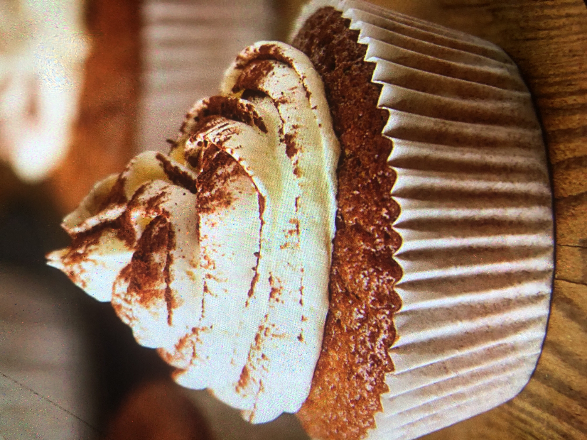 A close-up shot focuses on a single cupcake with white frosting and a dusting of cocoa powder. The cupcake is presented in a white paper liner and rests on a wooden surface. The background is blurred, showing hints of other cupcakes or baked goods. There are no people or animals visible in the image. The scene does not provide any information that would allow for the determination of its location in Tashkent, Uzbekistan, or any other city.