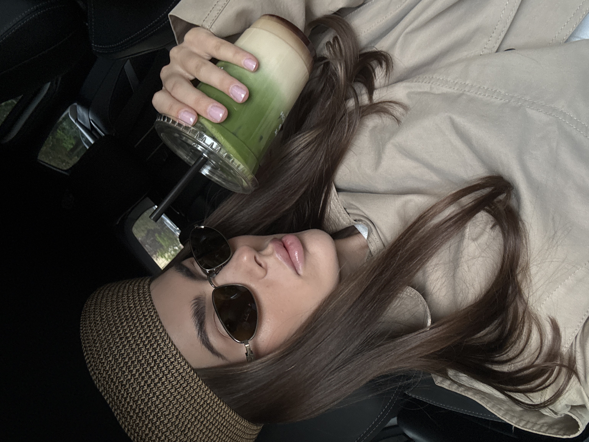 A woman with brown hair, wearing sunglasses and a brown hat, reclines in a black car seat, holding a clear plastic cup with a green matcha-colored beverage and a black straw. She wears a beige jacket and has manicured fingernails. The setting appears to be inside a car, potentially parked, given the angle and background. The specific location in Warsaw, Poland cannot be determined from the image.