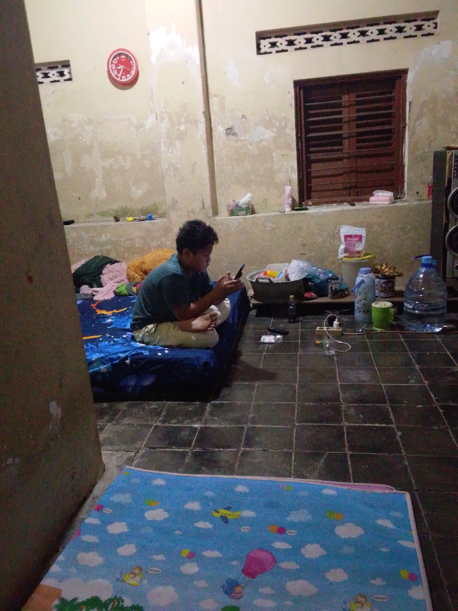 The image depicts an indoor scene, likely a modest living space, in Wirun, Indonesia. A young male is seated on a blue mattress on the floor, engrossed in his mobile phone. He is wearing a dark green t-shirt and light-colored pants. The room has a tiled floor and light-colored, somewhat weathered walls.
On a low shelf along the wall, there are various items including a red circular clock, a wooden-slatted window with horizontal blinds, a speaker, a large plastic water dispenser, a green mug, a cylindrical container with a "BLACK" label, and several bottles and bags. A plastic basin filled with items is also visible on the shelf. In the foreground, a blue mat with a cartoon pattern of clouds, airplanes, and hot air balloons is laid out on the floor. Electrical outlets and a power strip with a connected wire are present on the floor near the basin. The overall impression is that of a functional, if somewhat cluttered, domestic environment.