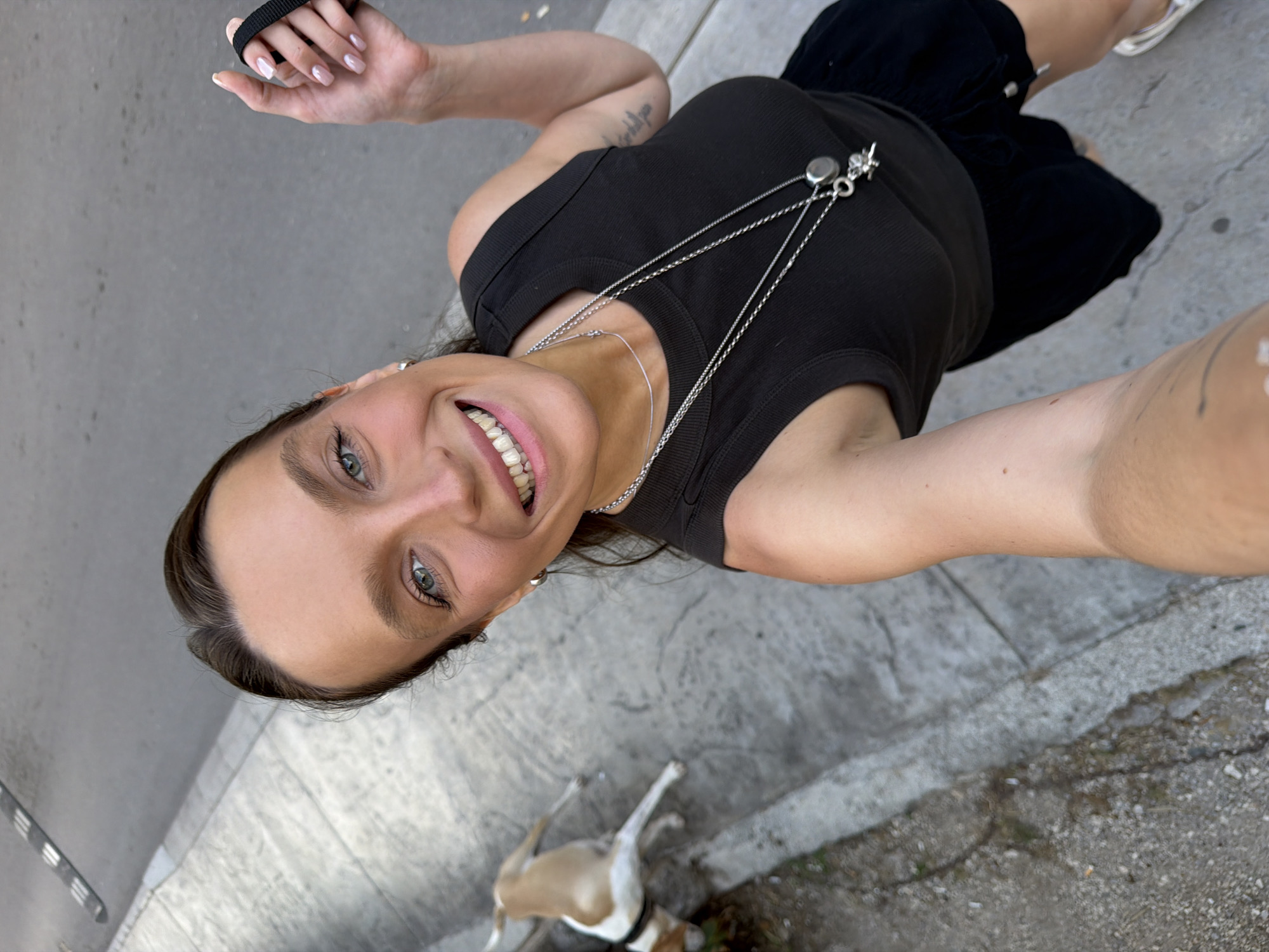 A woman with light brown hair and blue eyes smiles at the camera, taking a selfie. She is wearing a black tank top and black shorts, with a silver necklace. Her left hand is holding a black leash. To her left, a tan and white dog is on the ground, looking away from the camera. They are on a sidewalk in what appears to be a city, with a grey street in the background. The scene is likely Budva, Montenegro.