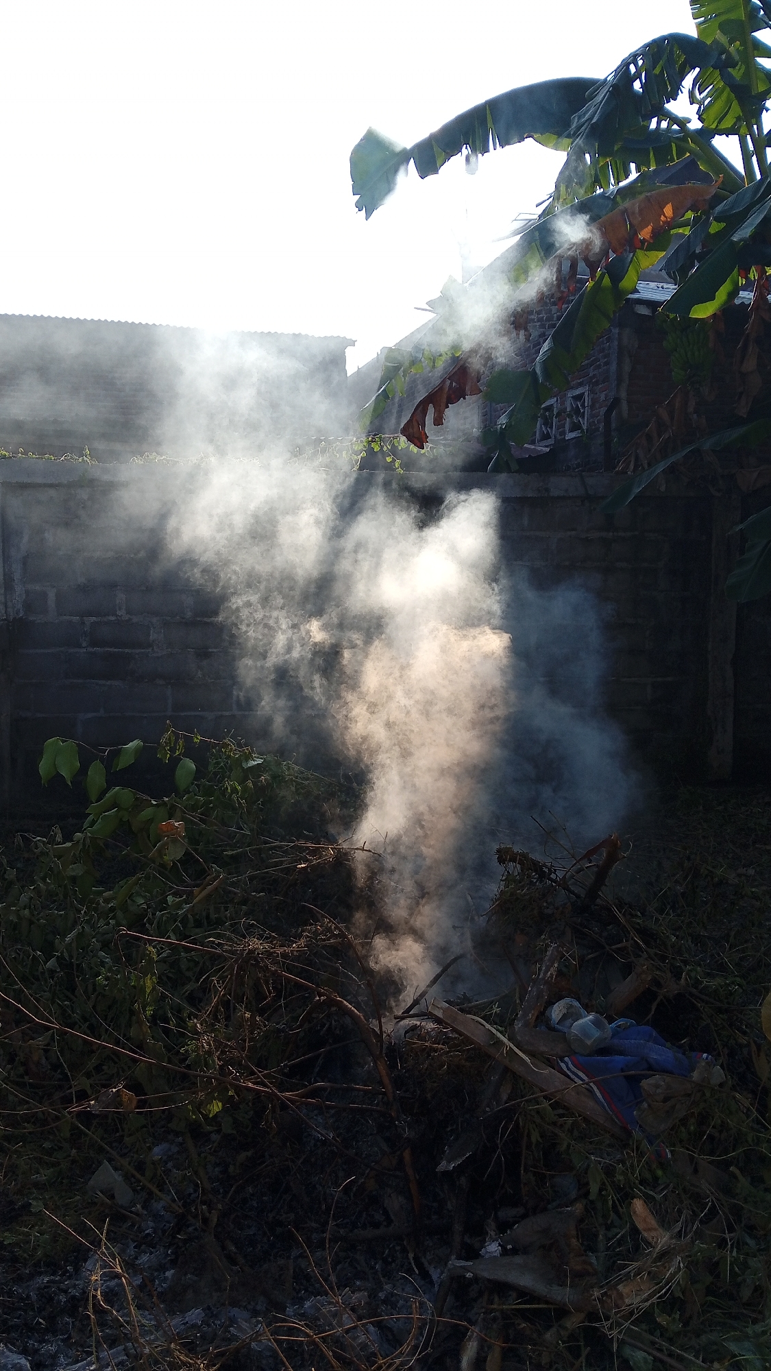 The image depicts an outdoor scene in Geulanggang Baro, Indonesia, with a pile of burning vegetation producing a significant amount of smoke. In the foreground, dry leaves, twigs, and branches form the fuel for the fire, with some evidence of ash. A blue garment with red and white stripes is visible amongst the debris. Behind the fire, a brick wall extends across the frame, topped with some greenery. In the background, several buildings with corrugated roofs are partially obscured by the smoke and bright sunlight. Banana leaves and trees are prominent in the upper right portion of the image, with sunlight filtering through them. The overall impression is of an outdoor burning activity under a bright sky.
