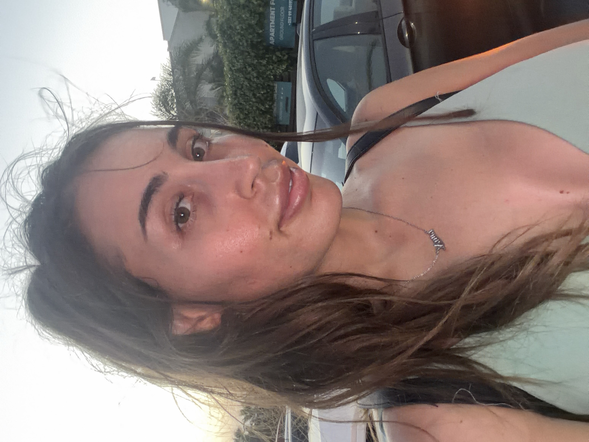 In Pervolia, Cyprus, a young woman with long, brown, windswept hair and brown eyes takes a selfie outdoors. She is wearing a light blue top and a silver necklace that spells out "Anna." Her skin is flushed, suggesting she may have been in the sun. In the background, palm trees and a building with a sign that reads "Apartment For Sale" are visible, along with parked cars. The overall lighting suggests it is late afternoon or early evening.