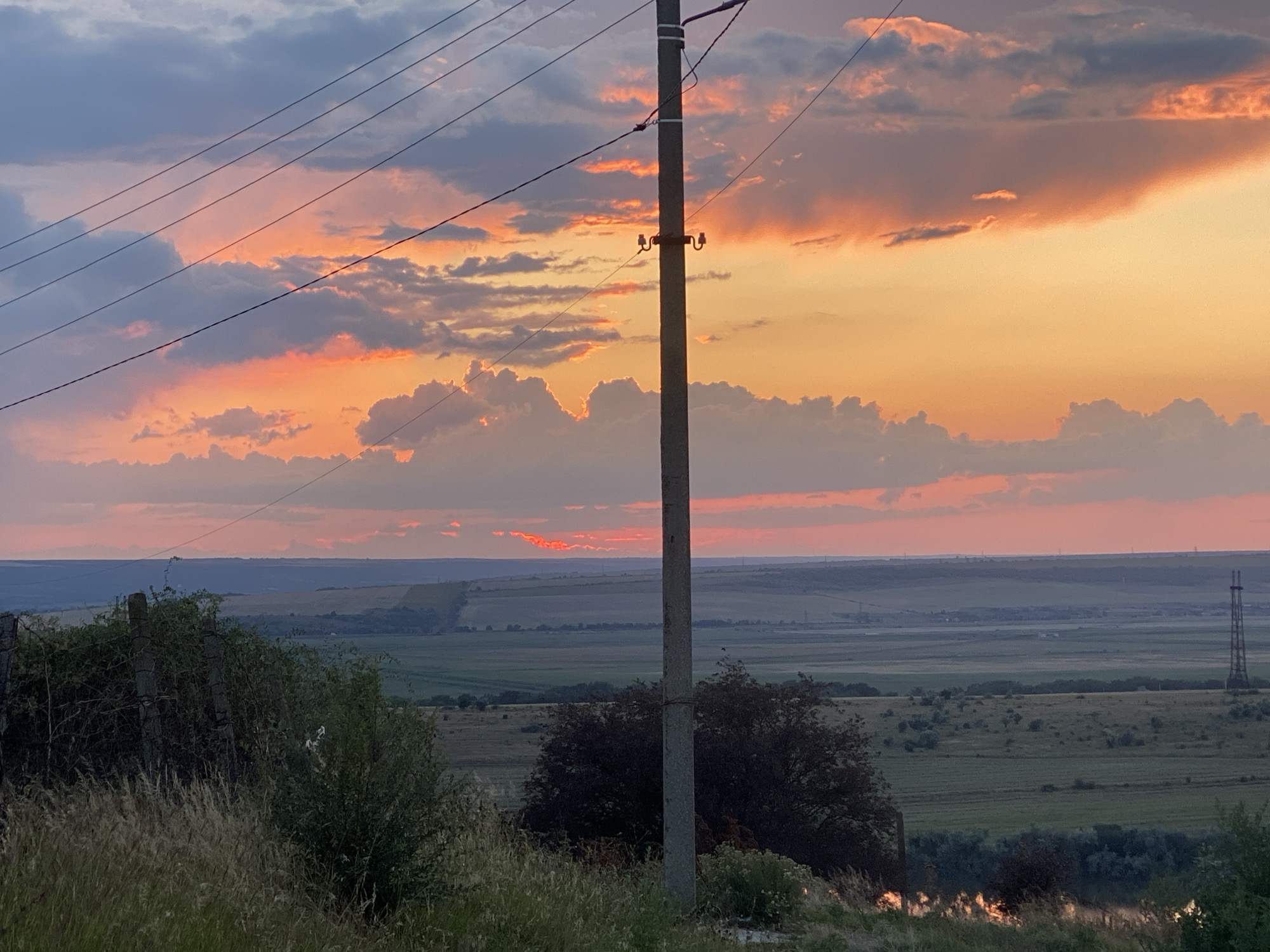 This is a landscape photograph taken in Cairaclia, Moldova, during sunset. The sky is filled with dramatic clouds in shades of orange, pink, and gray, illuminated by the setting sun. A prominent concrete utility pole with wires crosses the frame from left to right, positioned slightly off-center to the right. In the foreground, tall dry grass and foliage are visible, with some greenery and darker bushes. The midground consists of rolling hills and agricultural fields, appearing as a patchwork of green and brown. In the distance, a tall metal tower stands on the right side of the frame, and a sliver of water, likely a river or lake, reflects the sunset colors in the lower right corner. The overall scene captures a rural twilight ambiance with natural elements and man-made infrastructure. There are no people or animals visible in the image.