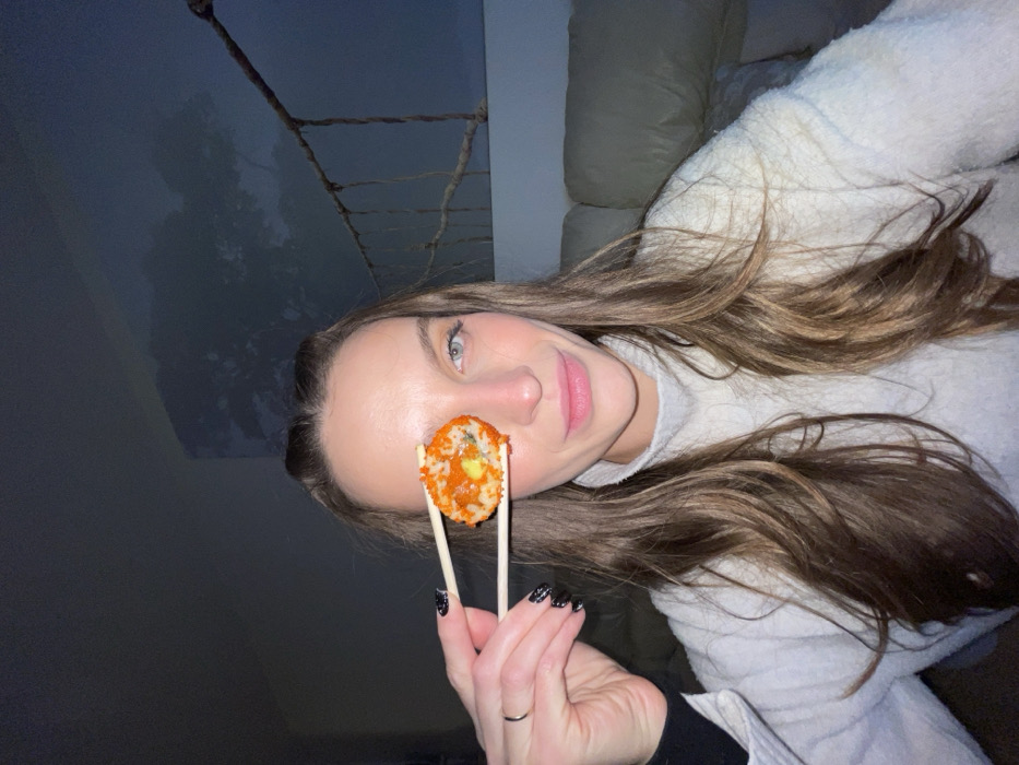 A woman poses with a sushi roll held up by chopsticks to cover her eye. She's wearing a fluffy, light-colored sweater and has long brown hair. The background is split between a dark grey wall and a soft, beige couch. She has glittery nail polish on and a ring on her left ring finger.