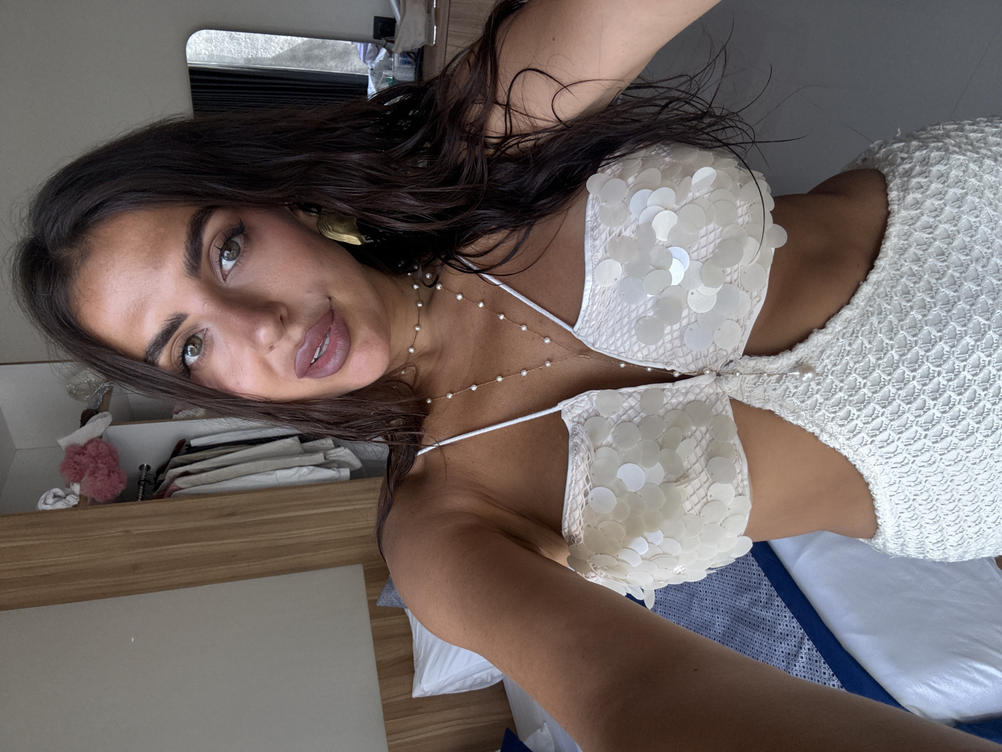 A medium-shot photograph shows a woman posing in what appears to be an interior space. The woman has dark hair, green eyes, and is wearing a white, sequined bikini top and a white crocheted bottom. She is also wearing a delicate necklace with pearls. The woman is looking at the camera with a slight smile.
The background reveals elements of a bedroom or living space, with a bed visible on the right side of the frame, covered with white sheets and a patterned blue and white blanket. A wooden shelf or cabinet is to the left, with items like folded clothes and a pink hair accessory. Overhead, there is a blurred view of what appears to be a sink area, adding to the impression of a domestic interior. The image has a slightly soft focus, contributing to a casual, personal feel.
The location in the city of Kuta Selatan, Indonesia, is not directly discernible from the image itself.