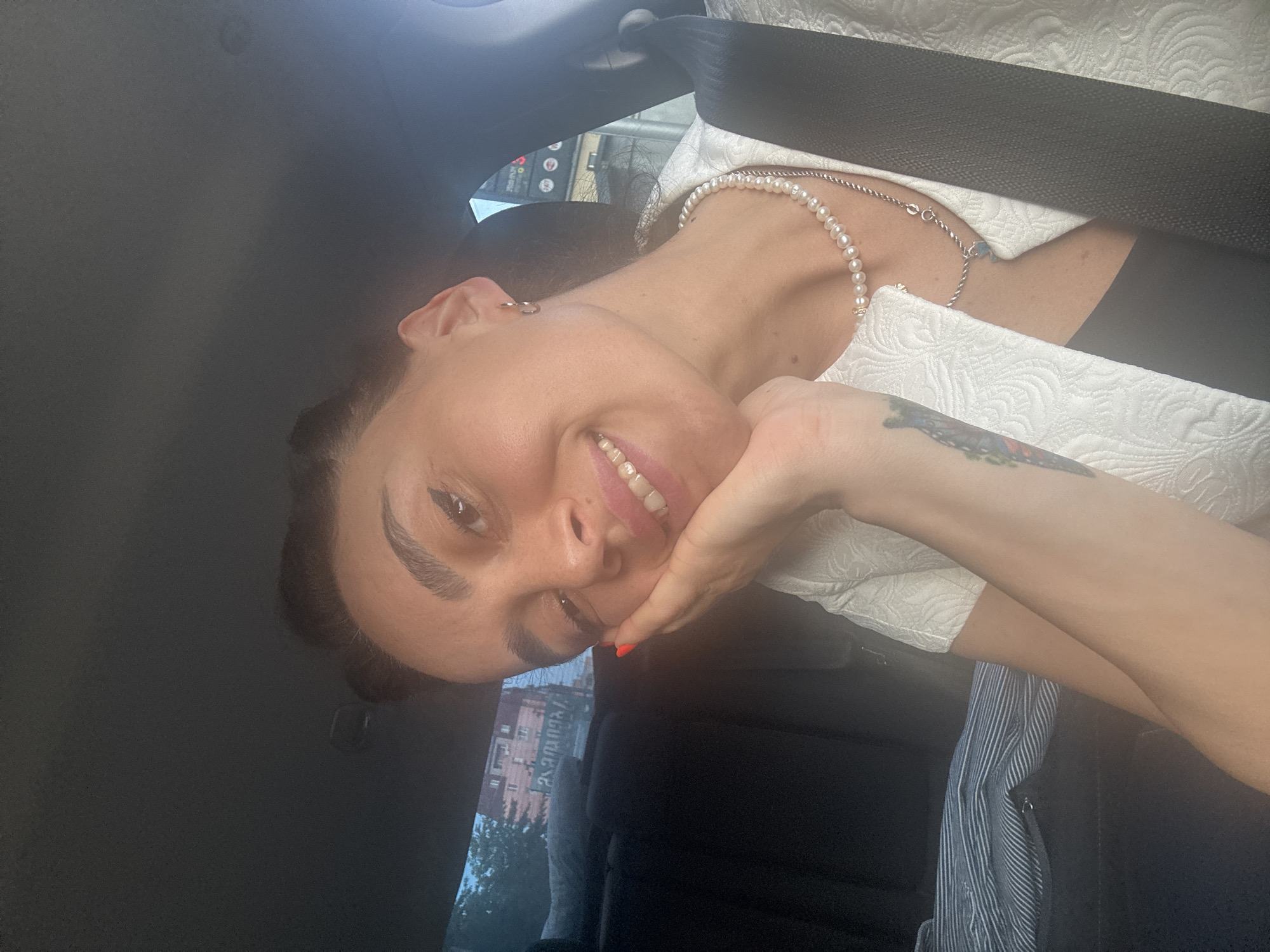 In this close-up selfie, a woman with dark hair pulled back is seated in the passenger seat of a car. She rests her right hand on her cheek, her eyes looking towards the camera with a slight smile. Her makeup includes winged eyeliner and bright lipstick. She is wearing a white textured jacket over a black top, with a pearl necklace and a silver chain with a pendant. A tattoo is visible on her left forearm. Through the car window, a blurry urban background is visible, suggesting the car is on a city street, possibly in Tbilisi, Georgia, given the architectural style and potential signage. The interior of the car is dimly lit, with a seatbelt visible across her chest.