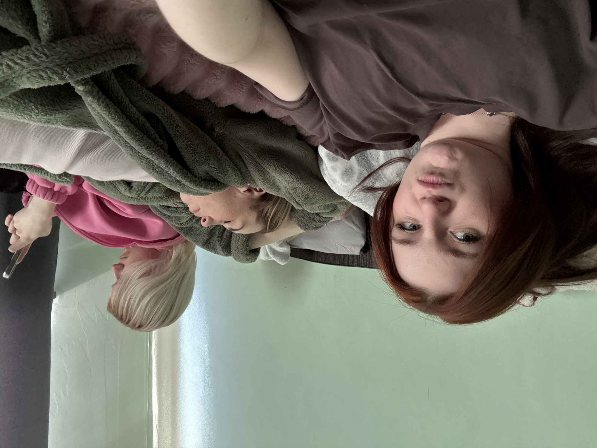 Three individuals are lying on a bed in what appears to be an indoor setting. The person closest to the foreground has reddish-brown hair and is wearing a brown top. Another person, partially obscured by a green blanket, is lying next to them. The third person, positioned to the left, has blonde hair and is holding a smartphone. The background consists of a light green wall. The location is unspecified.