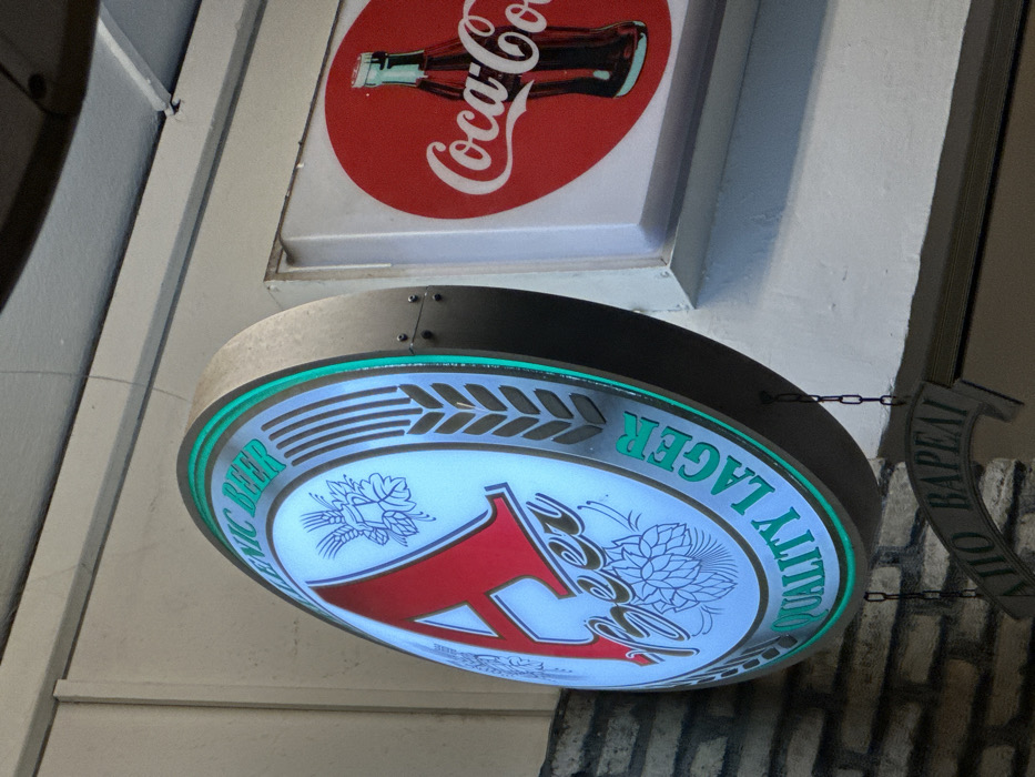 The image captures two illuminated commercial signs affixed to a building facade in Nea Erithrea, Greece, likely in the evening or at night. The upper sign is a rectangular Coca-Cola advertisement, displaying the classic red circular logo with white "Coca-Cola" script and a green glass bottle. Below it hangs a prominent, round, illuminated sign for a beer brand. This circular sign features a white glowing background adorned with detailed blue-grey illustrations of hops and barley. Green text curves along its edge; from an upright perspective, it reads "PREMIUM BEER" on the left side and "QUALITY LAGER" on the right. A large, stylized red letter, possibly "A", dominates the center, accompanied by the word "Beer" in a golden script. To the right of the round sign, a dark, shaped sign is partially visible, bearing white Greek text that reads "ΑΓΙΟ ΒΑΡΕΛ" (Holy Barrel), which confirms the Greek location context. The background consists of a light-colored wall and a section of decorative brickwork. No people or activities are present.