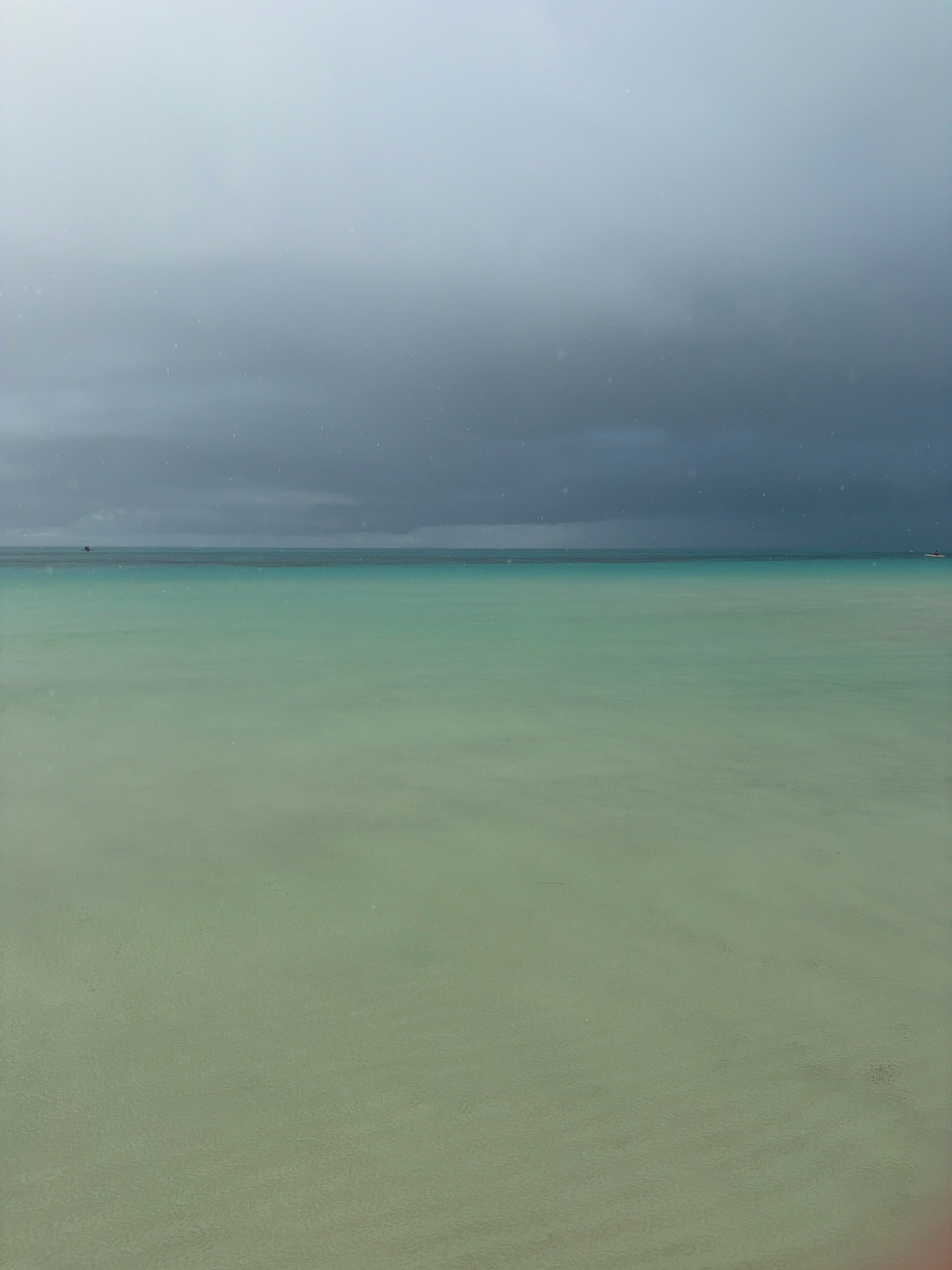 The image captures a serene seascape with a vast expanse of calm, shallow water in the foreground, transitioning to a deeper, darker turquoise horizon line. Above the water, a dramatic, overcast sky dominates, with heavy, dark gray clouds suggesting an impending storm or a recent rainfall. Small, white specks, possibly raindrops or sea spray, are visible against the dark sky and the water's surface. In the distance, a faint, dark silhouette of a boat is barely visible on the left side of the horizon. The overall scene is one of muted colors and a peaceful, yet somewhat somber, atmosphere, characteristic of Boracay Island's White Beach in Malay, Philippines, particularly during a cloudy day.
