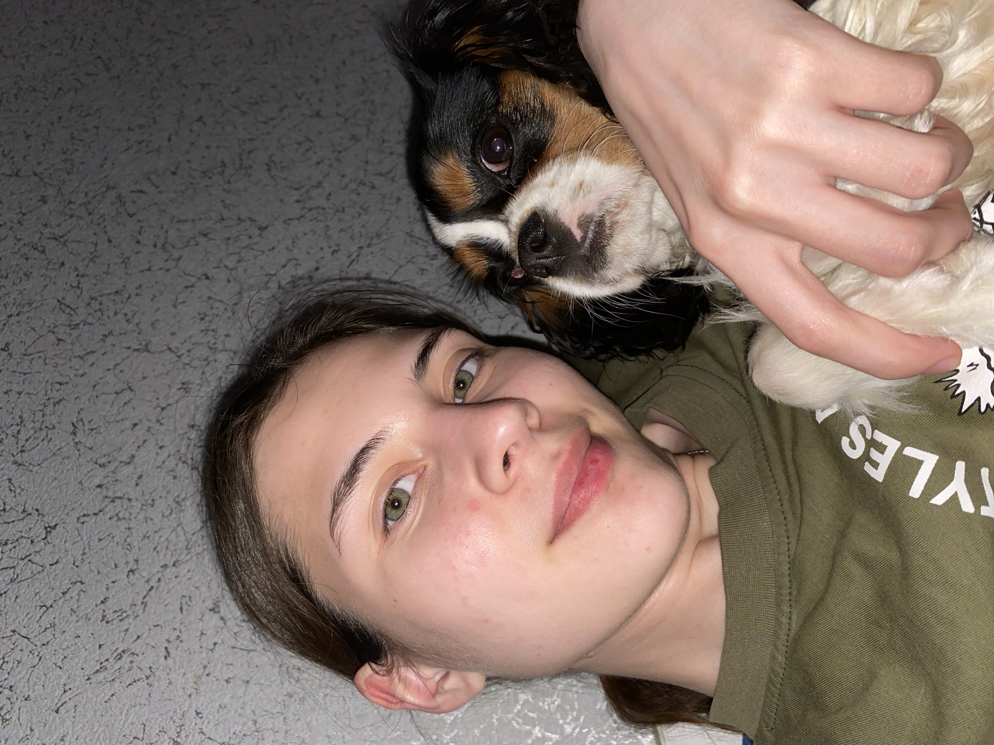 A person and a dog are visible in the image. The person is lying on a gray surface, possibly the floor, and is facing the camera. They have light skin, brown hair, and green eyes. The dog, a Cavalier King Charles Spaniel, is positioned above the person, its head close to the person's face. A hand is holding the dog. The person is wearing an olive-green T-shirt. The background is a textured gray surface. There is no indication of the location being Votkinsk, Russia.