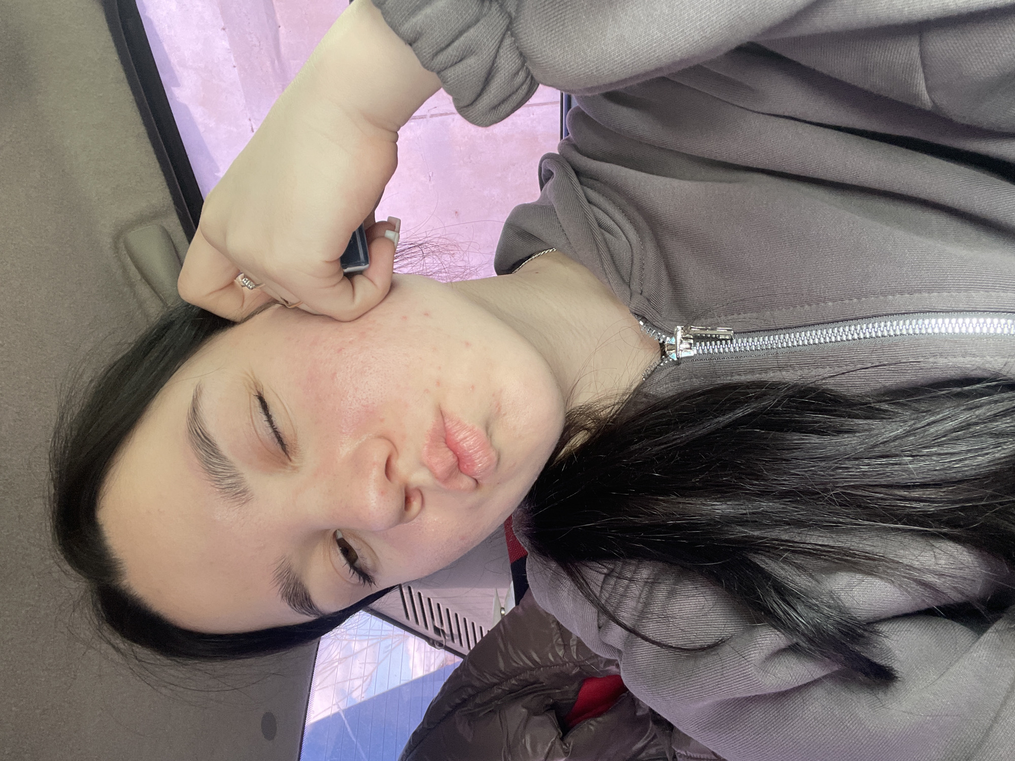 A woman is in the interior of a car. She is wearing a gray hooded sweatshirt with a zipper and has black hair. She is pouting her lips towards the camera. Her face has visible acne. She is holding a rectangular object near her face with her right hand. Her right hand has a ring on her finger and white nail polish. The car's interior includes a beige door panel and a reflective surface showing a window. The location cannot be determined to be the city of Узяр, Russia from this image.