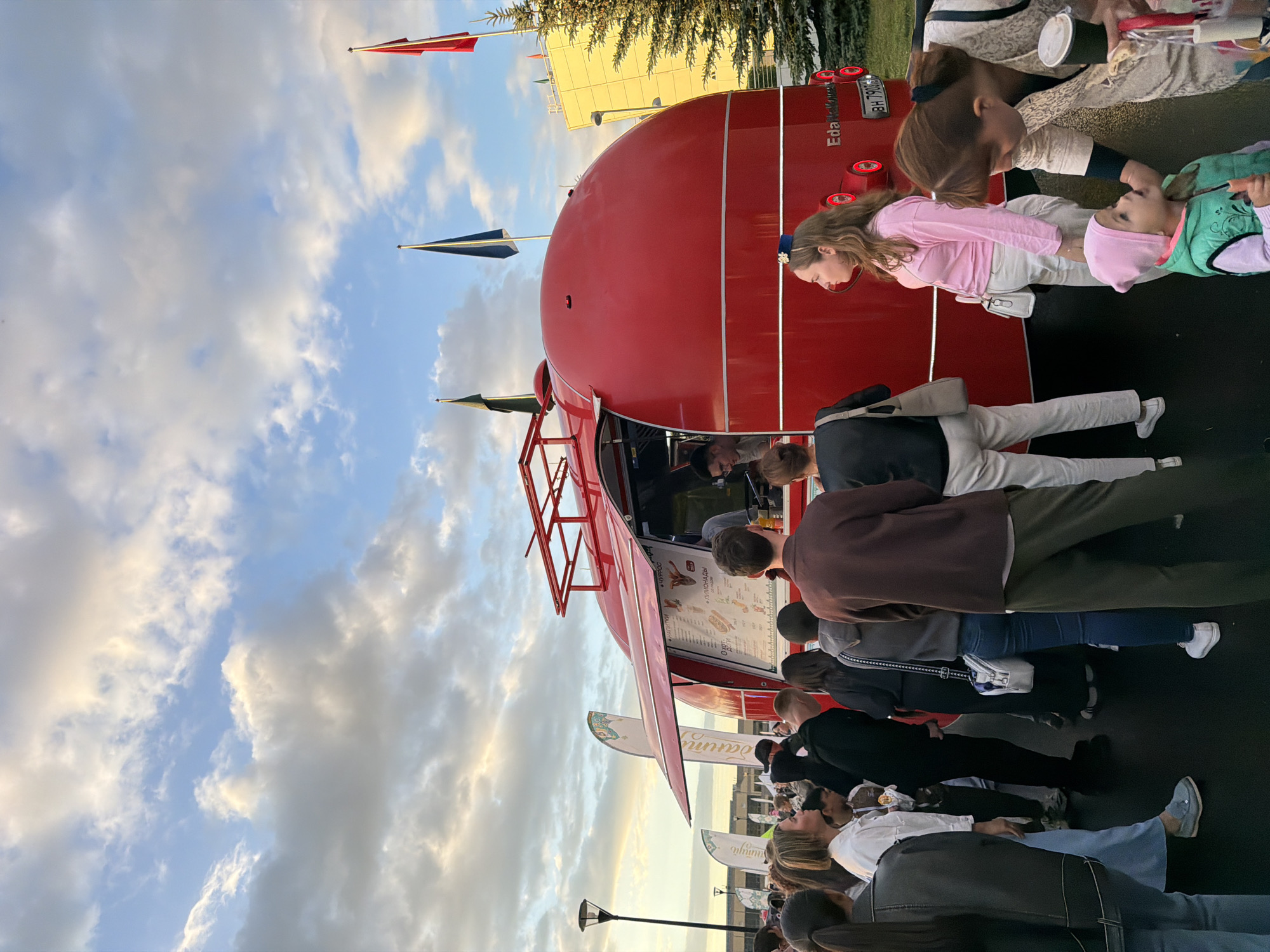 A large group of people stand in line outside a vibrant red, retro-style food truck. The truck is rounded and appears to be serving food. Some individuals are looking at the menu displayed on the side of the truck, while others are waiting patiently. The sky above is a mix of blue with scattered white and gray clouds, suggesting it could be late afternoon or early evening. Several flags are visible, two dark green and one red, adding to the festive atmosphere. In the background, a modern yellow building and a coniferous tree are present. A lamppost stands to the left of the scene. The ground appears to be paved.