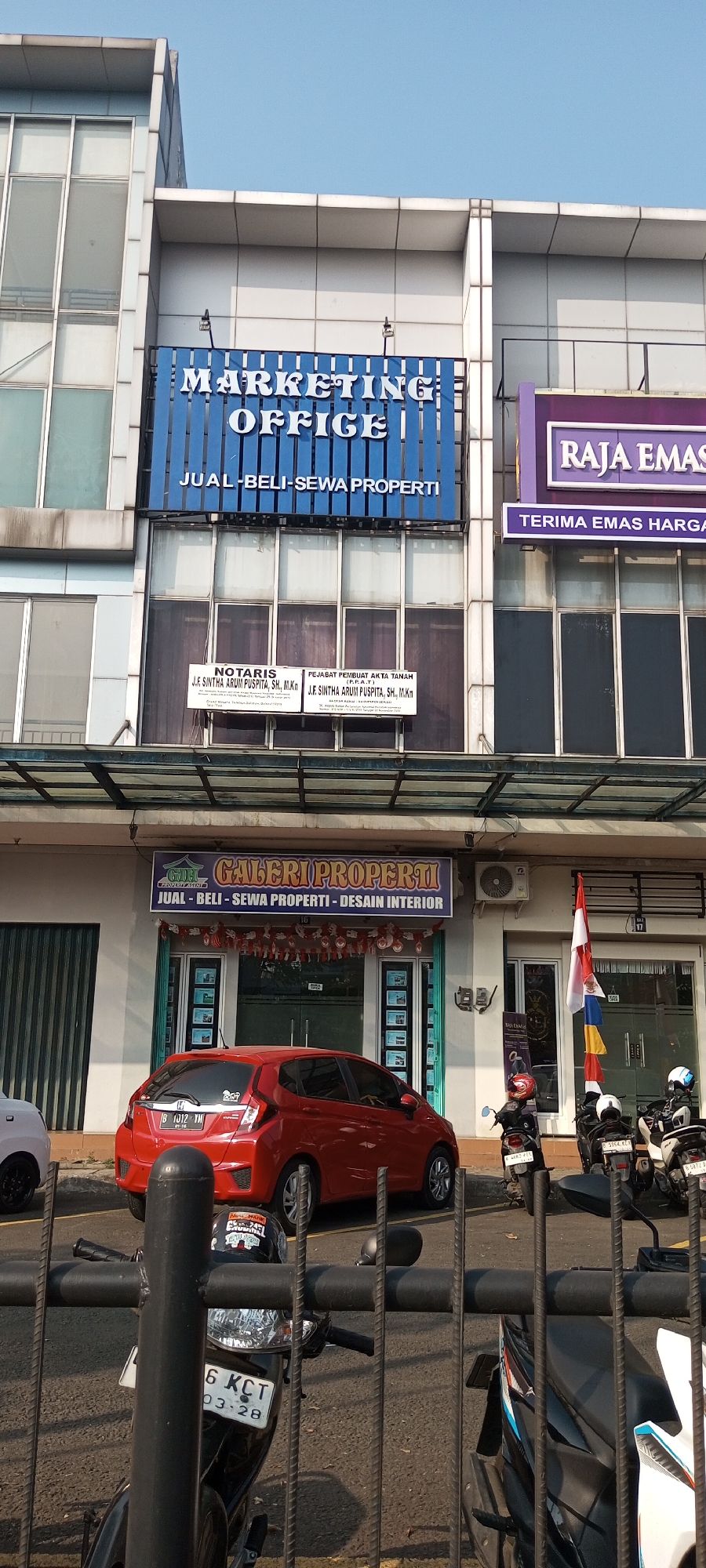 The image shows a street-level view of a commercial building in Lambangjaya, Indonesia. The building has multiple stories with large windows and visible signage. On the second floor, a blue sign reads "MARKETING OFFICE" with the subtitle "JUAL-BELI-SEWA PROPERTI". To the right of this sign, a purple sign reads "RAJA EMAS" with the subtitle "TERIMA EMAS HARGA". Below these signs, on the first floor, there are two white signs, one for "NOTARIS" and the other for "PEJABAT PEMBUAT AKTA TANAH (P.P.A.T.)".
In front of the building, parked cars and motorcycles are visible. A red Honda Jazz is prominently featured in the foreground. Several motorcycles are parked on the sidewalk. There are no people clearly visible in the image, but their presence is implied by the parked vehicles. The environment appears to be an urban street, with a clear sky overhead. The image is taken from a slightly low angle, looking up at the building.