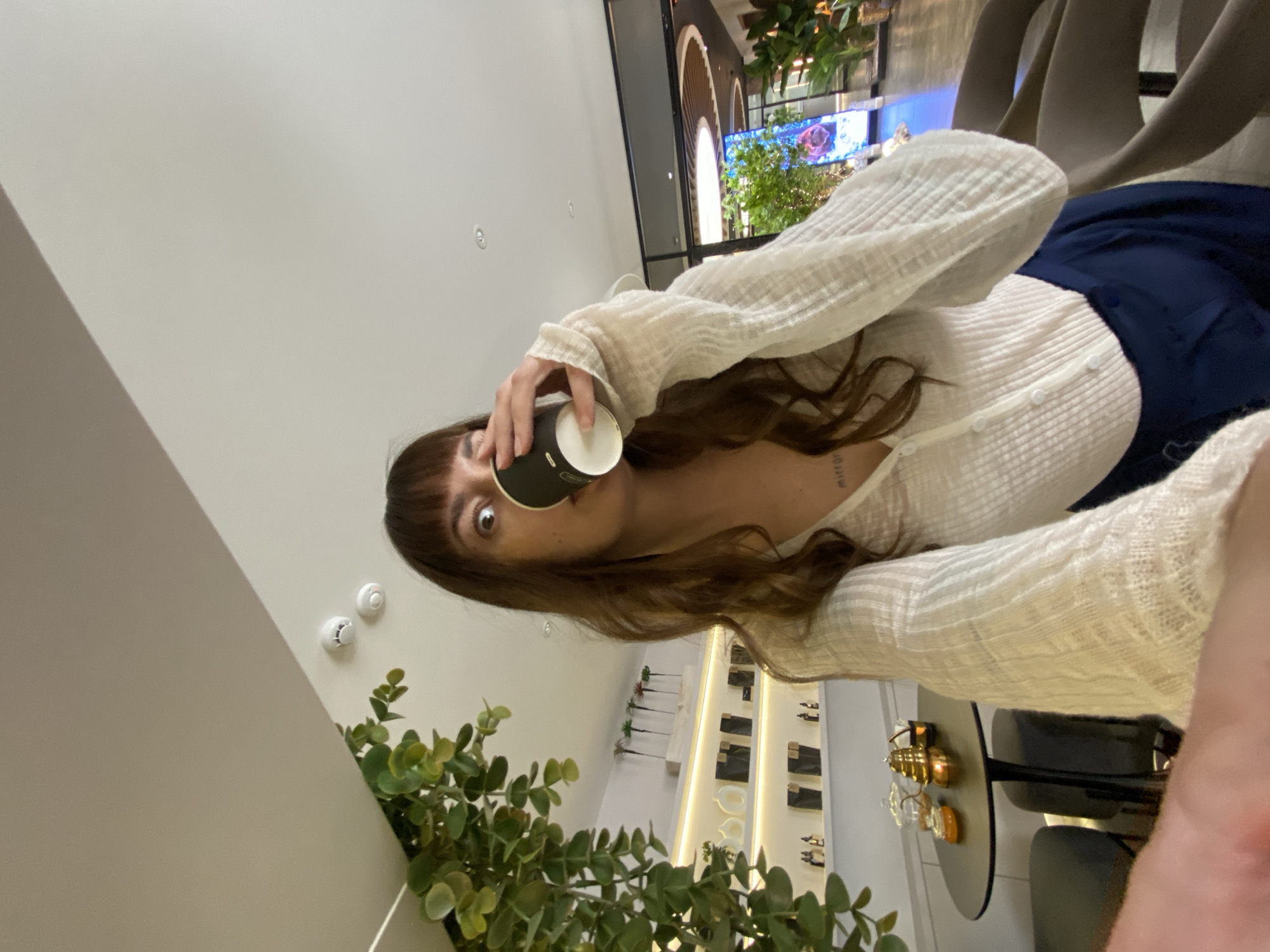 A young woman drinks from a black paper cup in what appears to be a public space or commercial establishment. She has brown hair with bangs and is wearing a white button-down shirt and dark blue pants. In the background, there are chairs, plants, and a display of merchandise. The location is not identifiable as Votkinsk, Russia, based on the image.