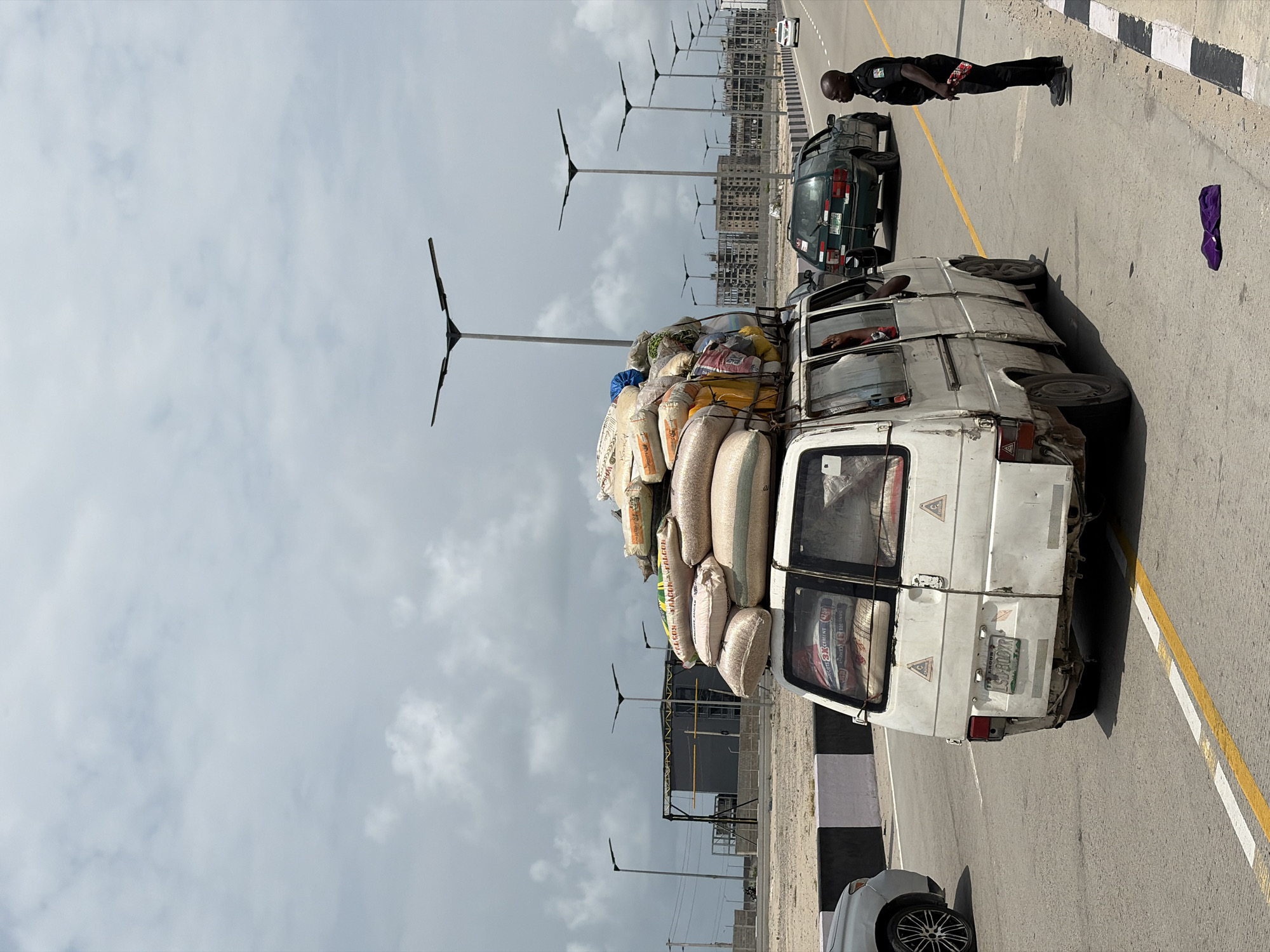 The image captures a scene in Alaguntan, Nigeria, featuring an overladen white van stopped on a paved road. The van is packed high with numerous sacks, obscuring much of the vehicle's upper structure. A uniformed officer stands near the van, interacting with the driver, whose arm is visible through the van's open window. A dark-colored car is parked closely behind the van. In the background, rows of streetlights line the road, with buildings visible in the distance under a cloudy sky. A purple object rests on the pavement. The van's license plate reads "LS 900 XG."