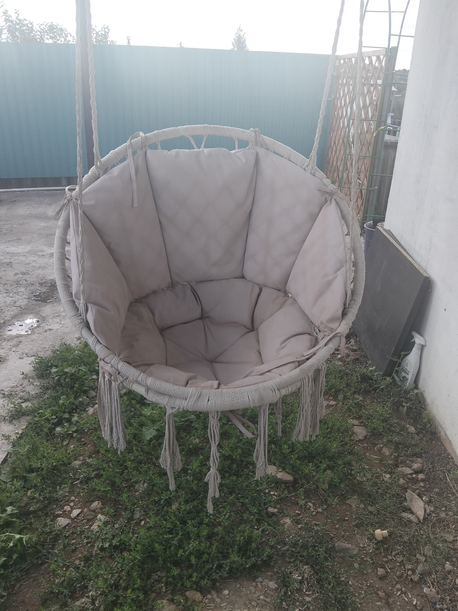 A beige hanging macrame chair with a plush cushion sits in an outdoor area. The chair is suspended by two thick ropes from an unseen support structure above. The chair has a circular frame and is adorned with decorative tassels. The background shows a teal corrugated metal fence, a wooden lattice fence, and a white wall of a building. The ground is covered with grass, stones, and some debris, including a puddle of water. A black rectangular object and a white spray bottle are leaning against the wall. The scene appears to be in the city of Oktyabrsky, Russia.