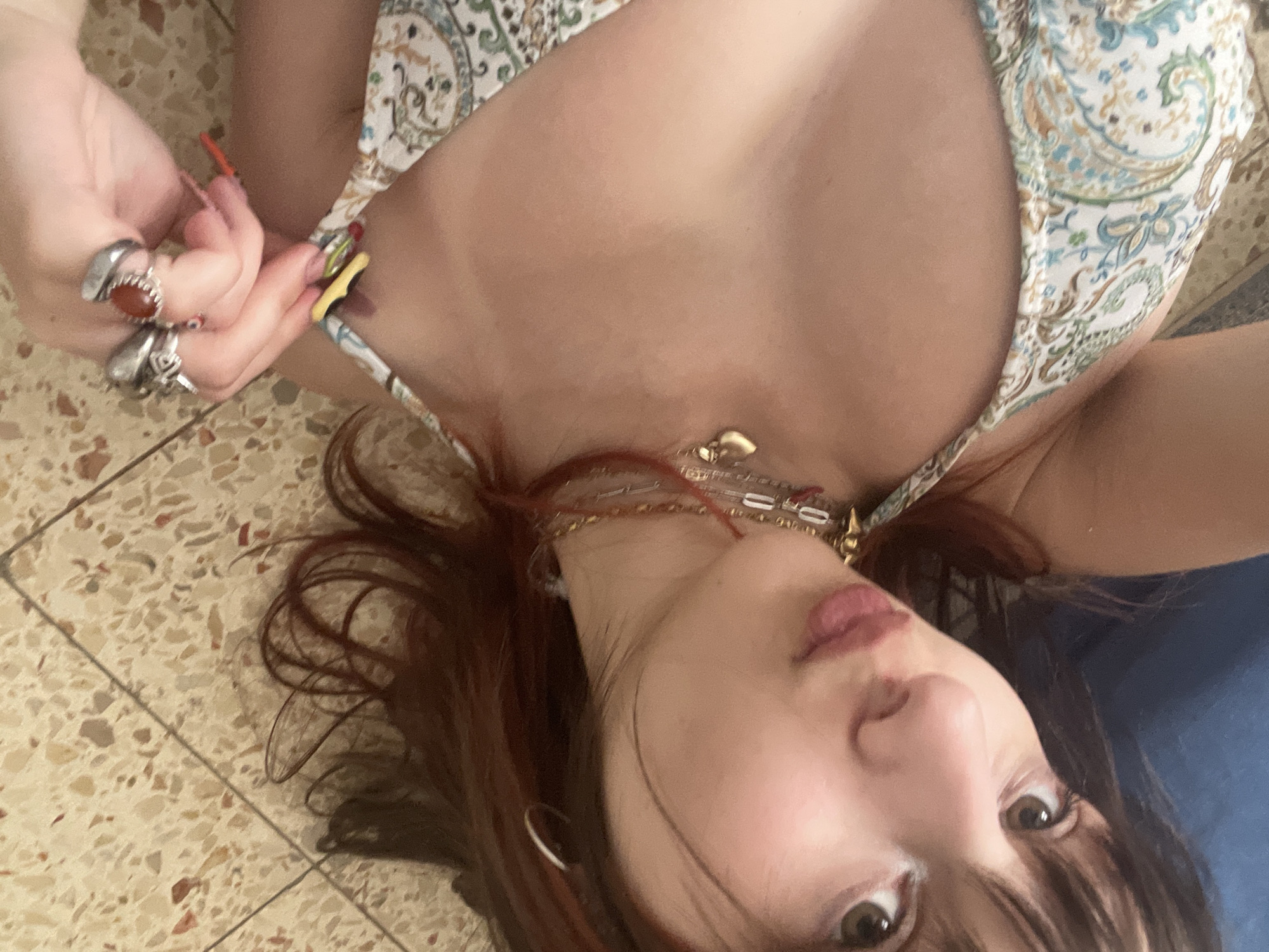 A woman is lying on a tiled floor. She has reddish-brown hair with bangs and is wearing multiple necklaces. She's wearing a paisley patterned top. Her left hand is visible, adorned with multiple rings, and holding a strap of her top. The backdrop includes the tiled floor and a blue object. The image was not taken in Petah Tikva, Israel.