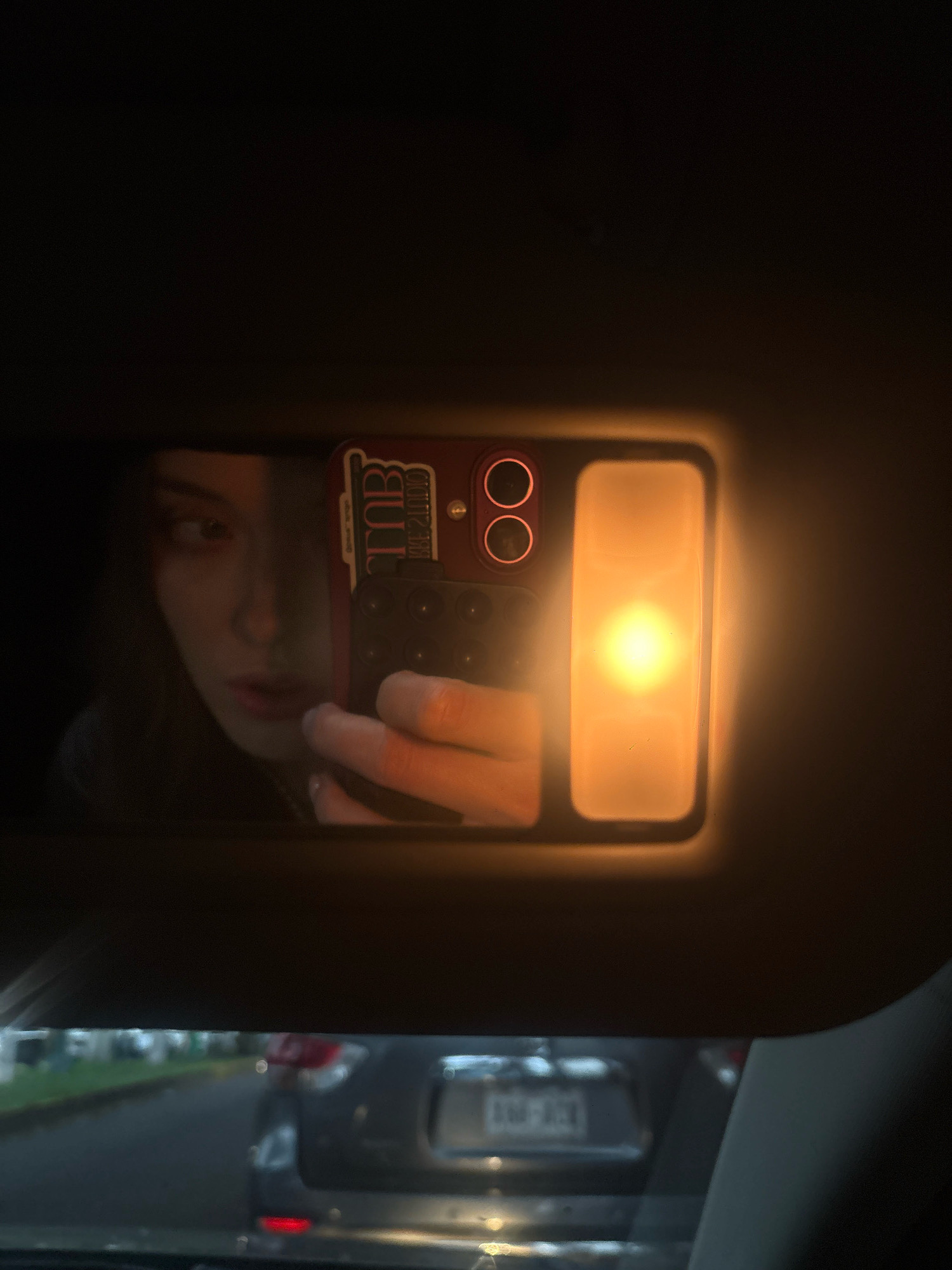 The image shows a reflection in a car's visor mirror. A young woman is visible, holding a smartphone to take the photo. Her face is partially obscured by shadow, and she is looking to her left. The smartphone has a red case with a design that appears to say "TNB KHESTUDIO". A bright, warm light emanates from a rectangular fixture next to the phone in the mirror. The background of the reflection shows the interior of a car, with a view of traffic outside, suggesting the car is on a street. The overall lighting is dim, with the primary light source being the car's vanity light. There is no discernible location in Mexico City, Mexico.
