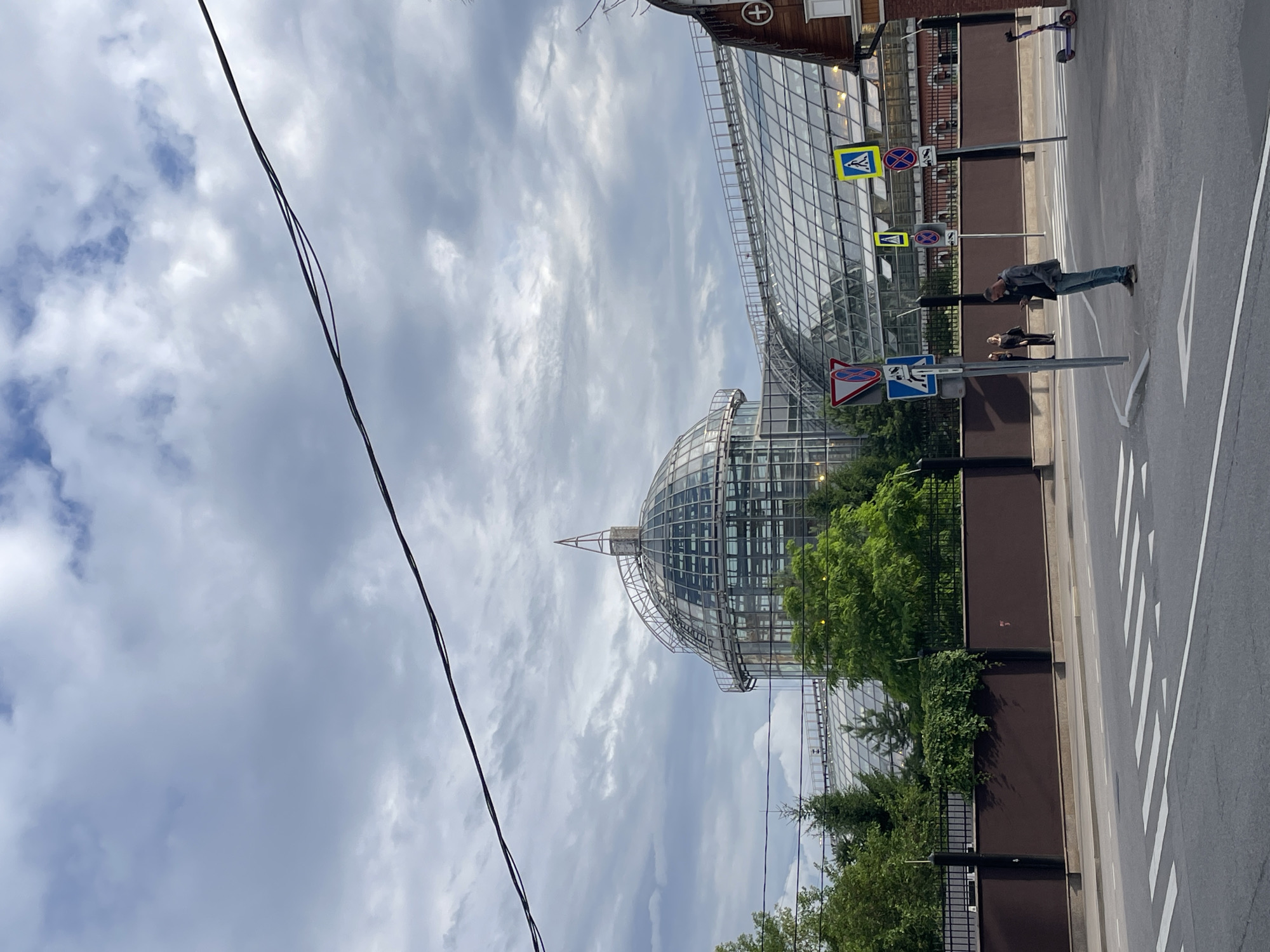 The image depicts a street scene in Moscow, Russia, with a prominent glass-domed building in the background. The building appears to be a modern structure with a geodesic dome as its centerpiece, suggesting it could be a botanical garden, exhibition center, or similar public facility. Green trees and fencing line the perimeter of the building, separating it from the road.
In the foreground, a street with white pedestrian crossing lines is visible. A few people are present: one person is walking on the sidewalk, and another is standing near a traffic sign. There is also a child or smaller person standing near the adult on the sidewalk. A purple electric scooter is parked on the sidewalk further down the street.
Several traffic signs are visible, including a pedestrian crossing sign and a "no entry" or "do not enter" sign. The weather appears overcast, with a cloudy sky, suggesting it might be a mild or cool day. The presence of power lines crossing the frame from the left indicates an urban environment. The overall impression is a snapshot of everyday life in a modern city, with a focus on architecture and urban infrastructure. No explicit text is clearly legible beyond the symbols on the traffic signs.