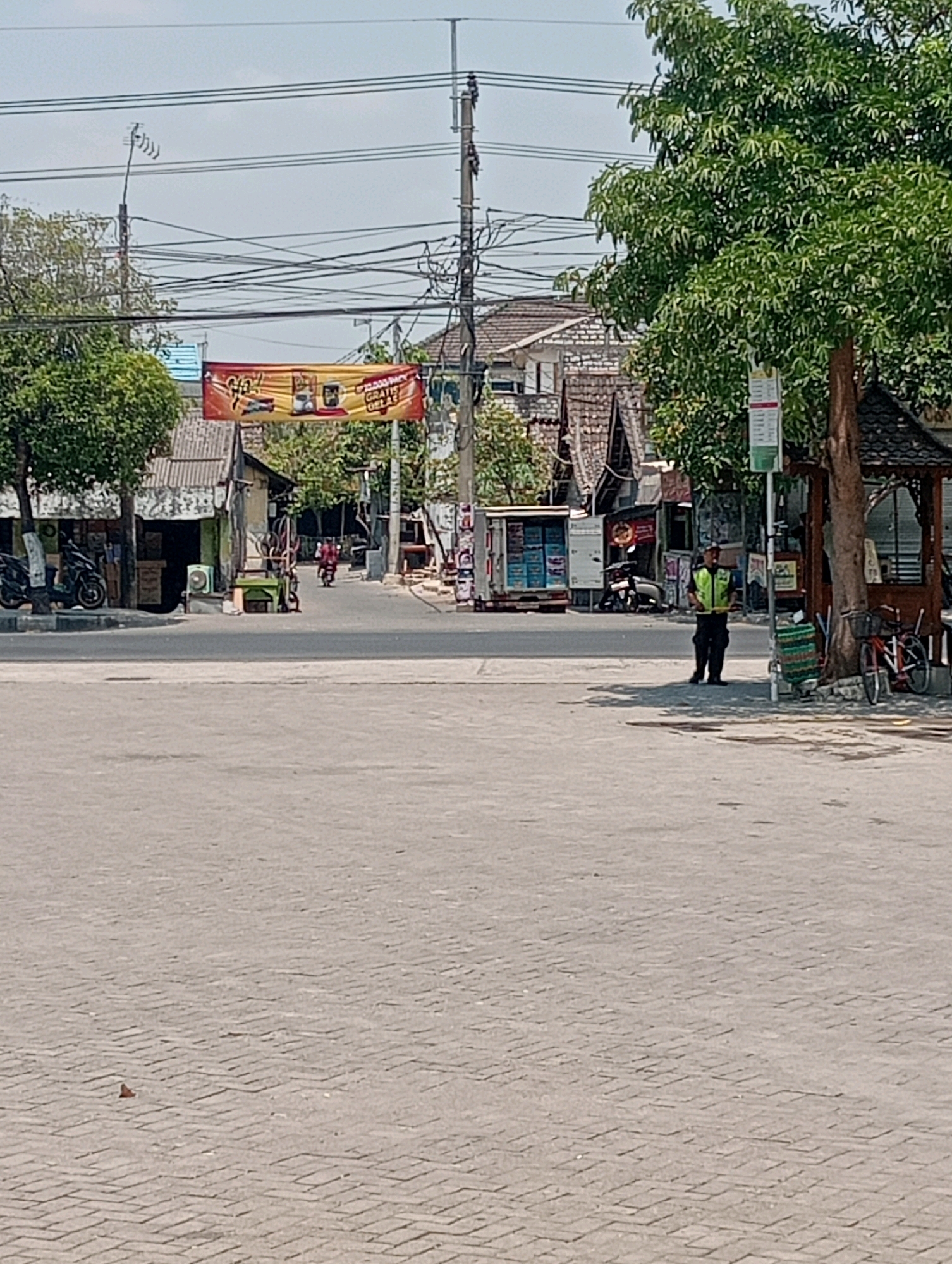 The image depicts a street scene in Soko, Indonesia, under a clear sky. In the foreground, a wide paved area with interlocking bricks stretches across the frame. To the right of the paved area, a man wearing a high-visibility yellow vest and dark pants stands near a wooden kiosk-like structure and a bicycle. Behind him, a mature tree provides shade, and a red bicycle is parked.
The middle ground shows a street with a few vehicles, including motorcycles parked along the left side and one being ridden further down the road. Various small shops and buildings line the street, some with corrugated metal roofs. A banner with red and yellow colors, featuring text that appears to be an advertisement, is strung across the road.
In the background, more buildings, trees, and utility poles with numerous wires are visible, contributing to a sense of urban infrastructure. The overall impression is a bustling, functional street in a tropical or subtropical environment.